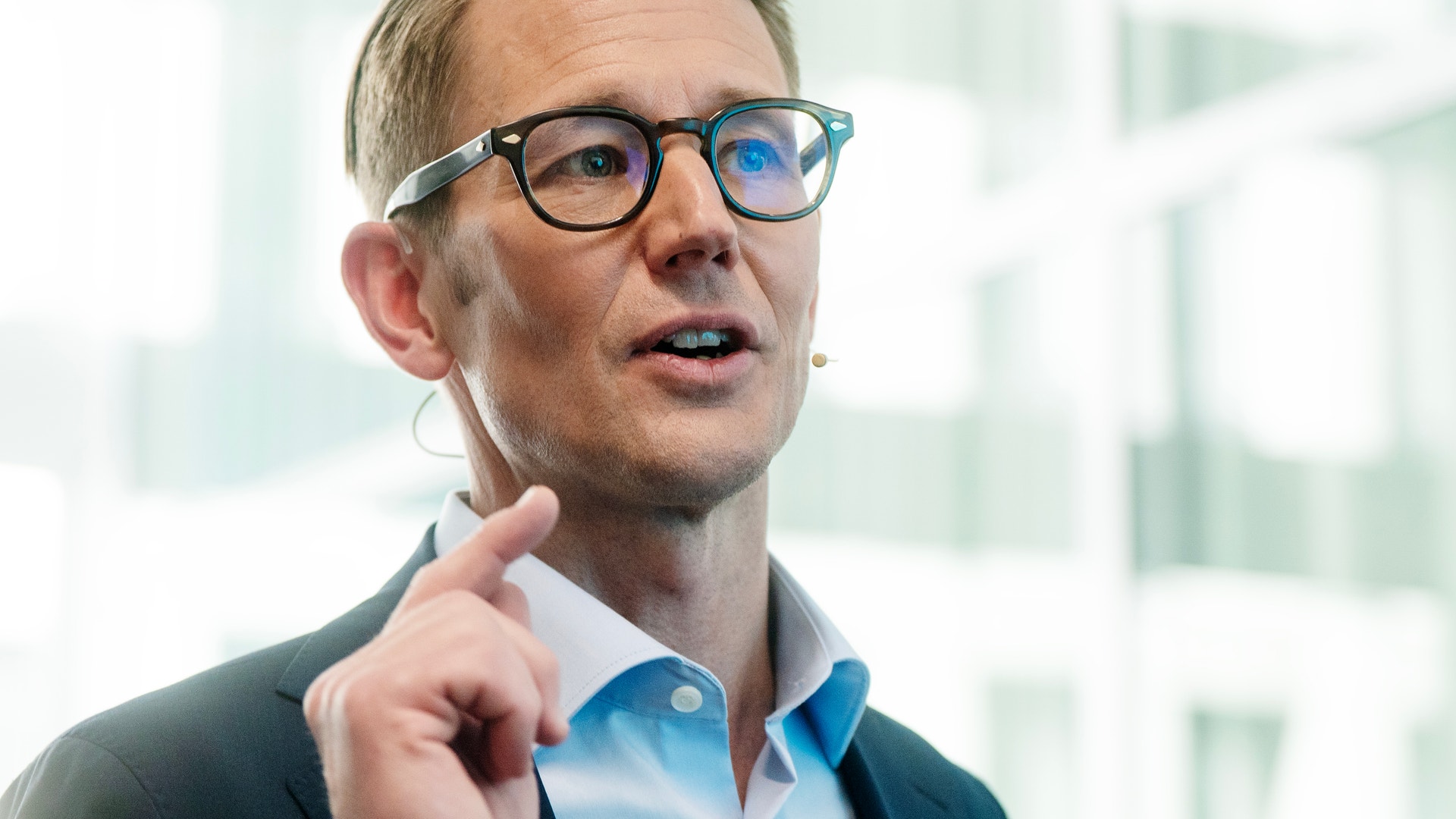 Man med glasögon i blå skjorta och kavaj som håller ett tal framför ett modernt kontorslandskap.