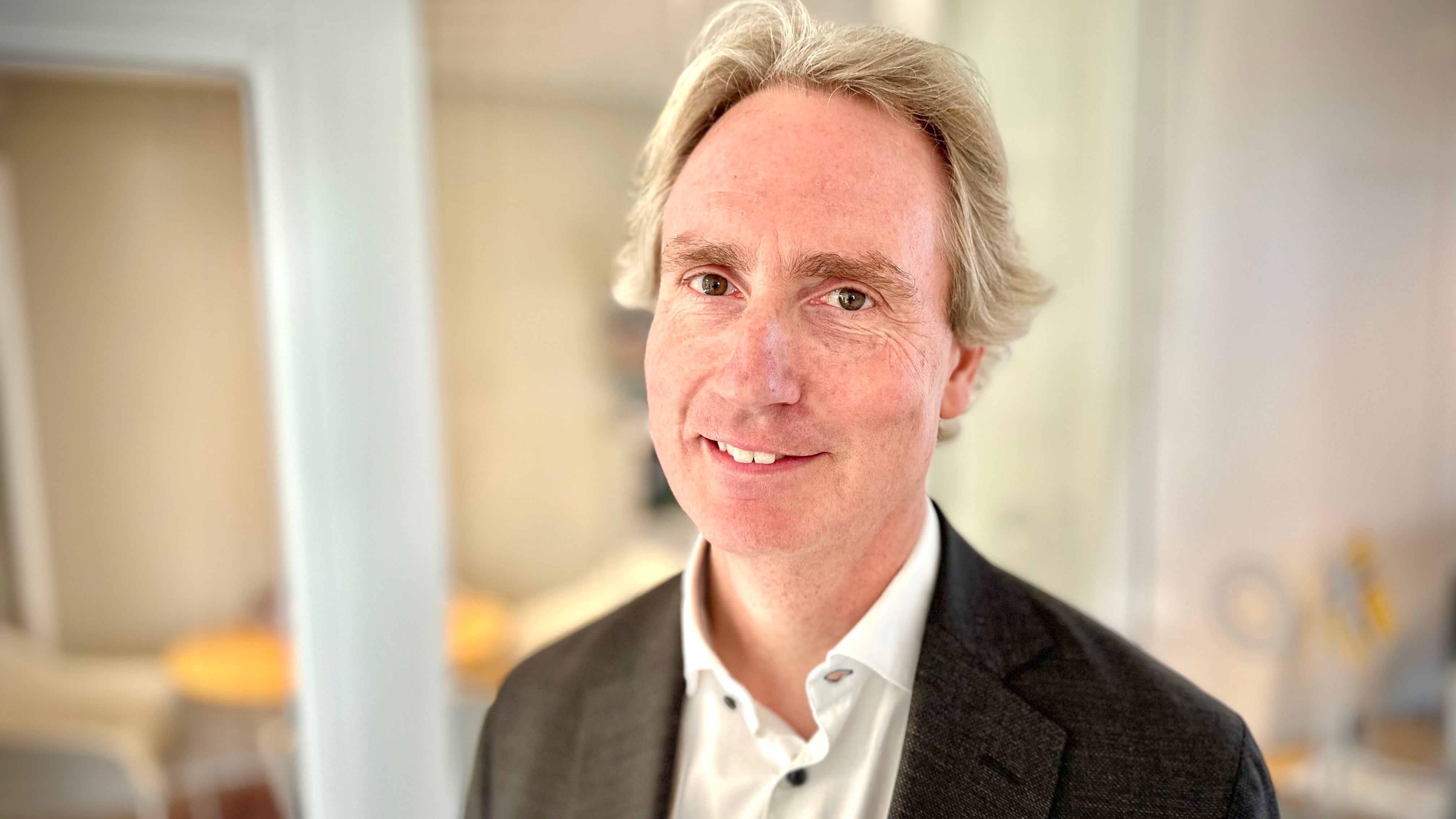 Porträtt av en man med blont hår, mörkgrå kavaj och vit skjorta, som ler mot kameran.