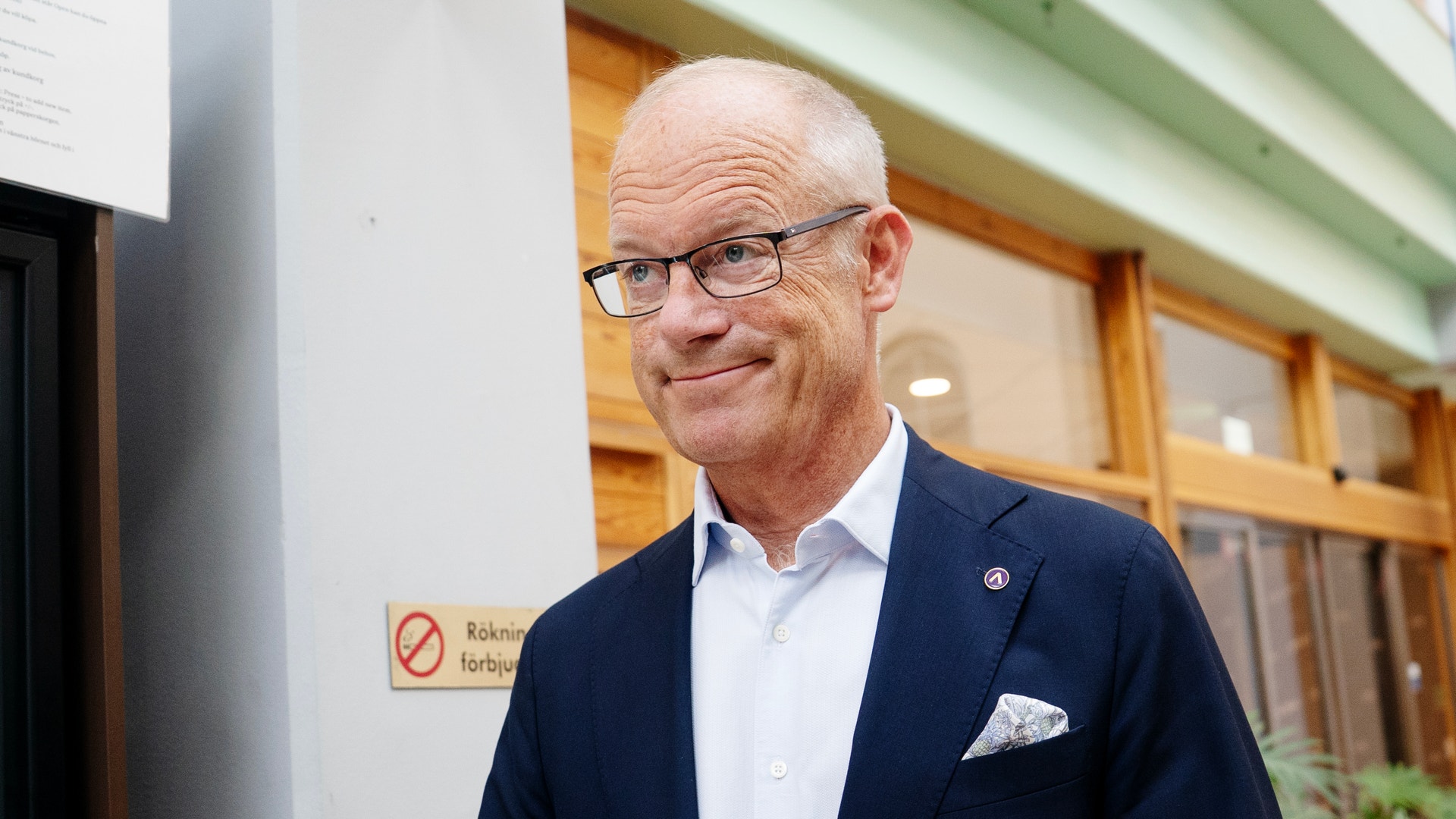 Porträtt av en man med glasögon som bär mörk blazer och ljus skjorta. Han står inomhus framför träfönster och ler mot kameran.
