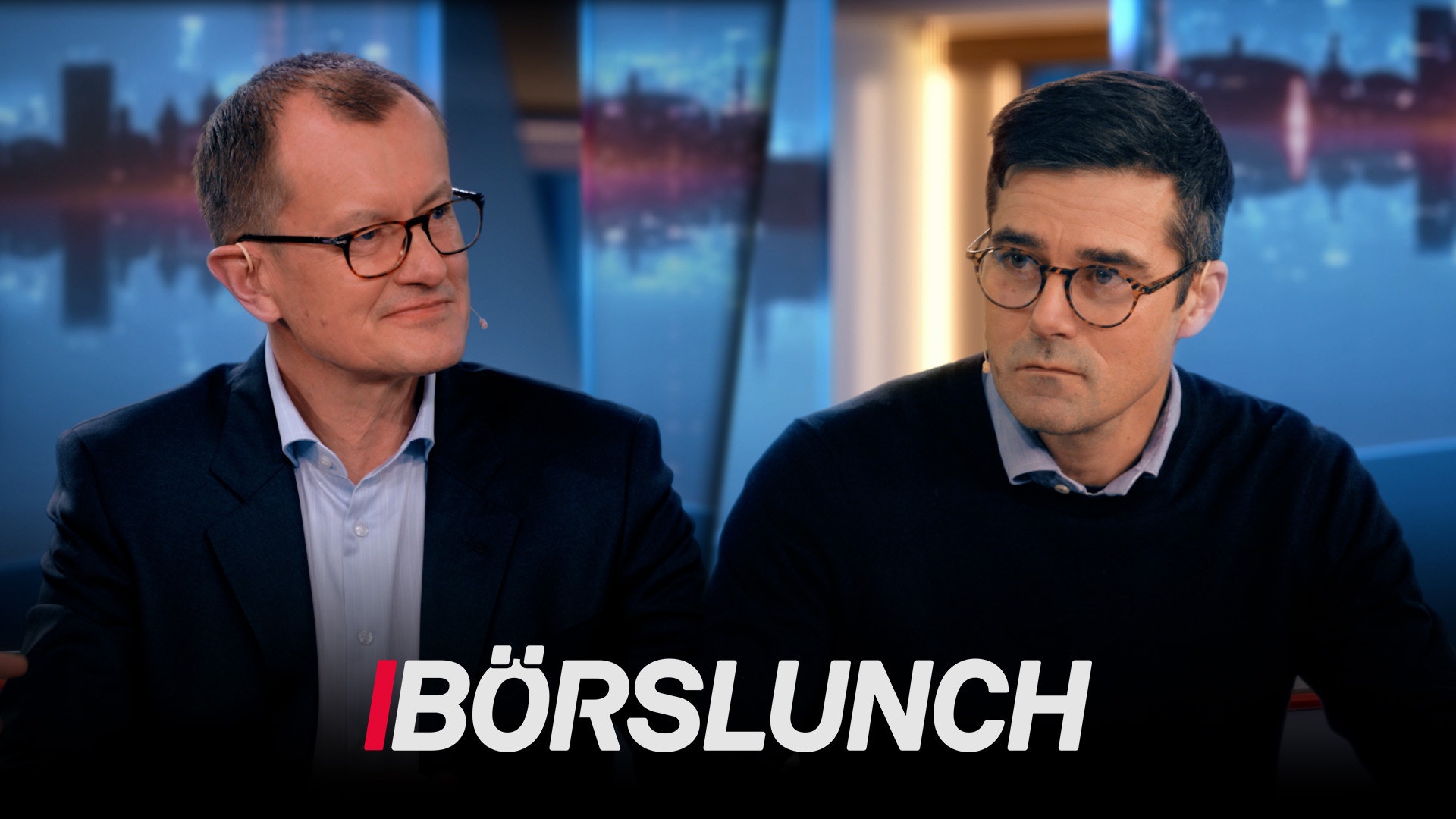 Två män i affärskläder diskuterar i en TV-studio med stadsvy i bakgrunden.