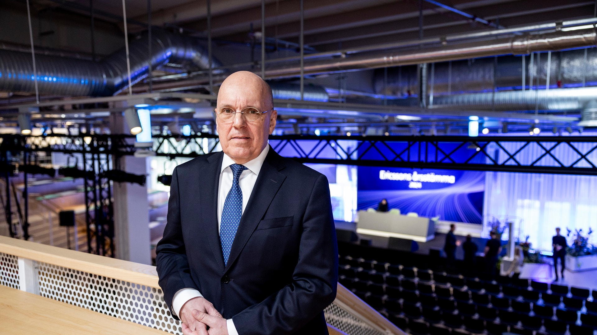 Man i mörkblå kostym och blå slips står vid räcke i modernt eventvenue med blå belysning i bakgrunden.