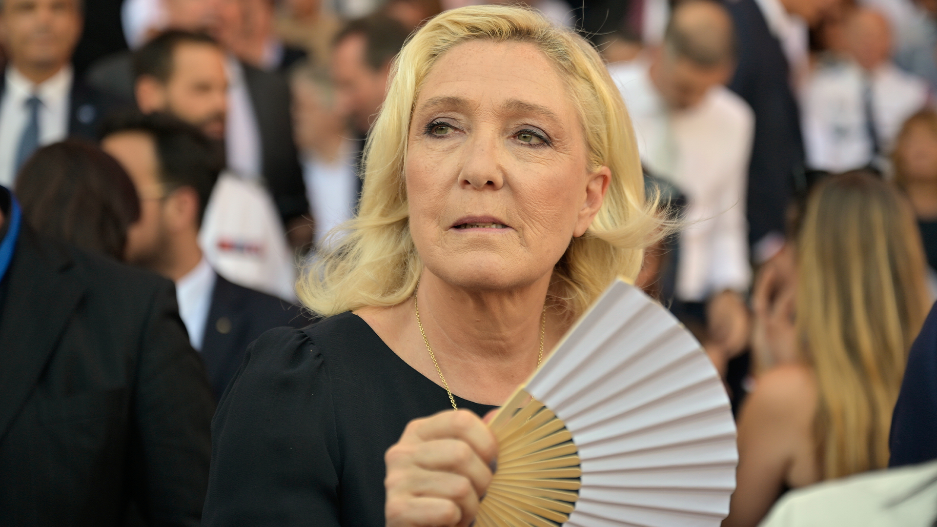 Marine Le Pen, en blond kvinna i svart klänning, står bland andra människor och håller en solfjäder.