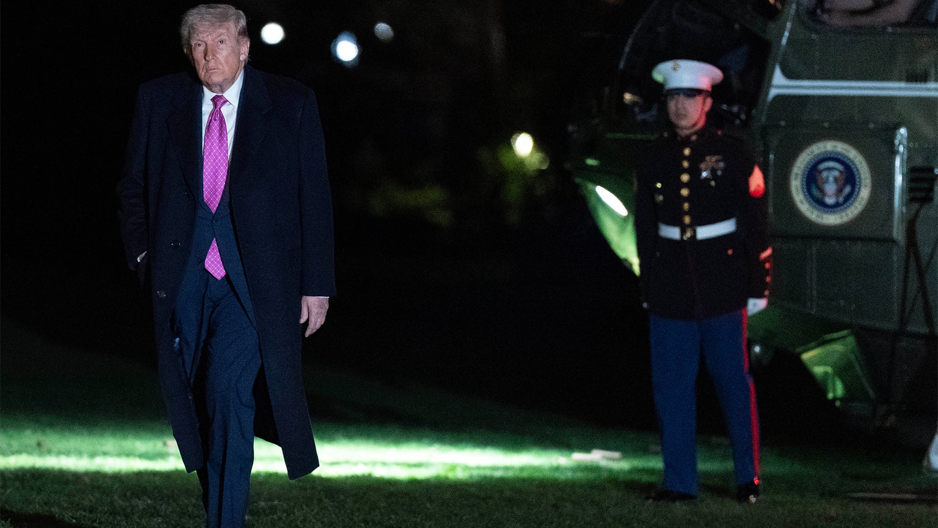 Man i mörk kostym går på upplyst gräsmatta på kvällen med militär personal och officiell fordon i bakgrunden.
