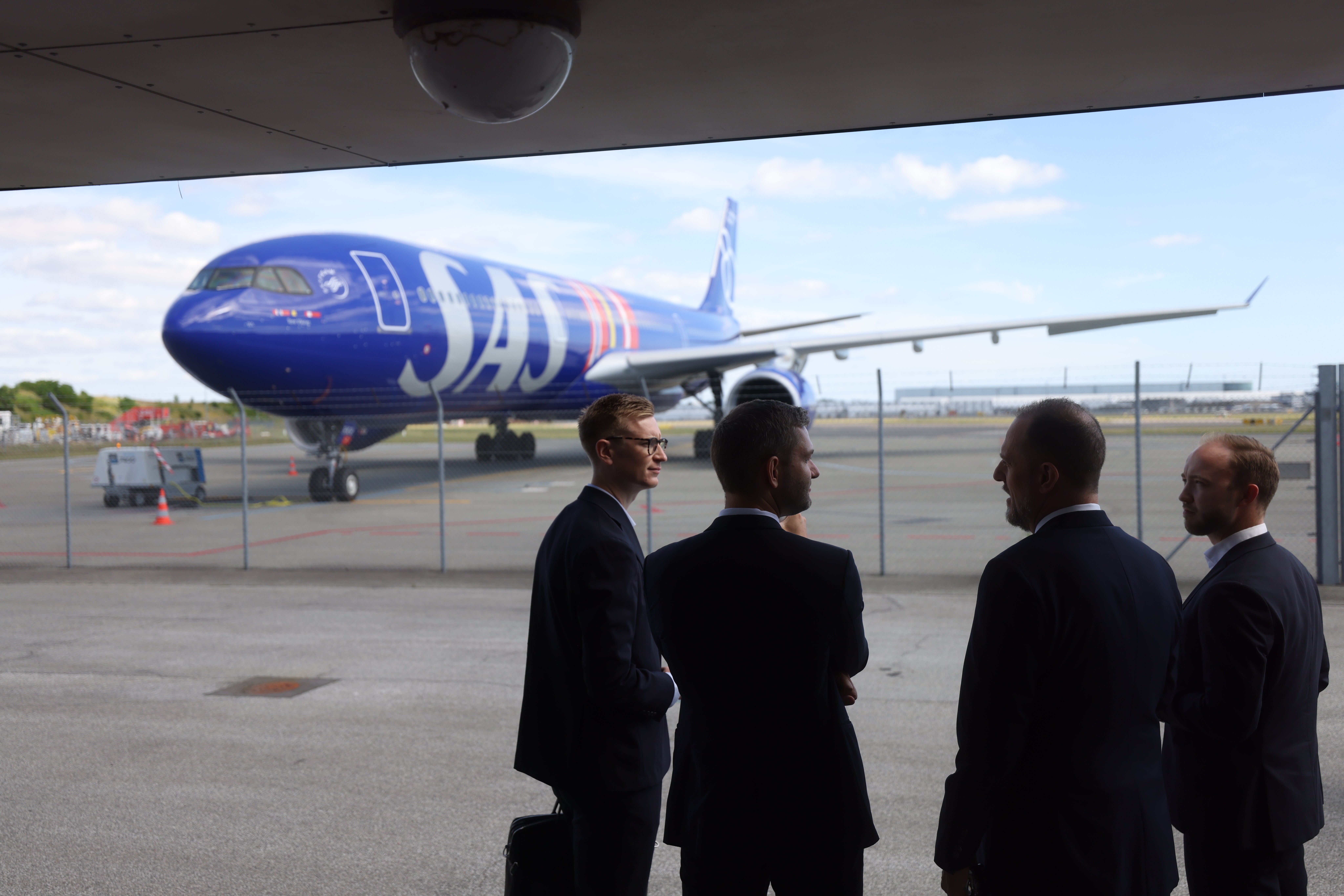 Fyra män i kostym står och samtalar på en flygplats, med ett SAS-flygplan synligt i bakgrunden.