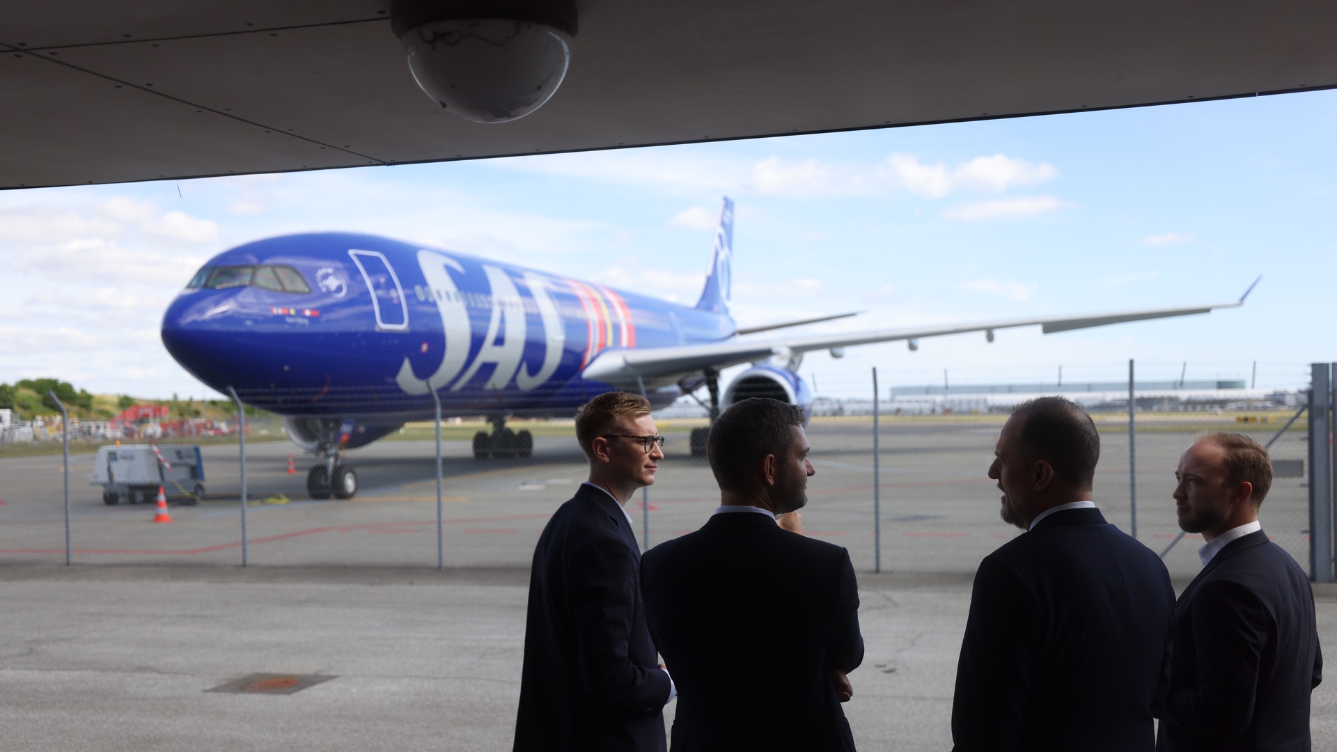 Fyra män i kostym står och samtalar på en flygplats, med ett SAS-flygplan synligt i bakgrunden.