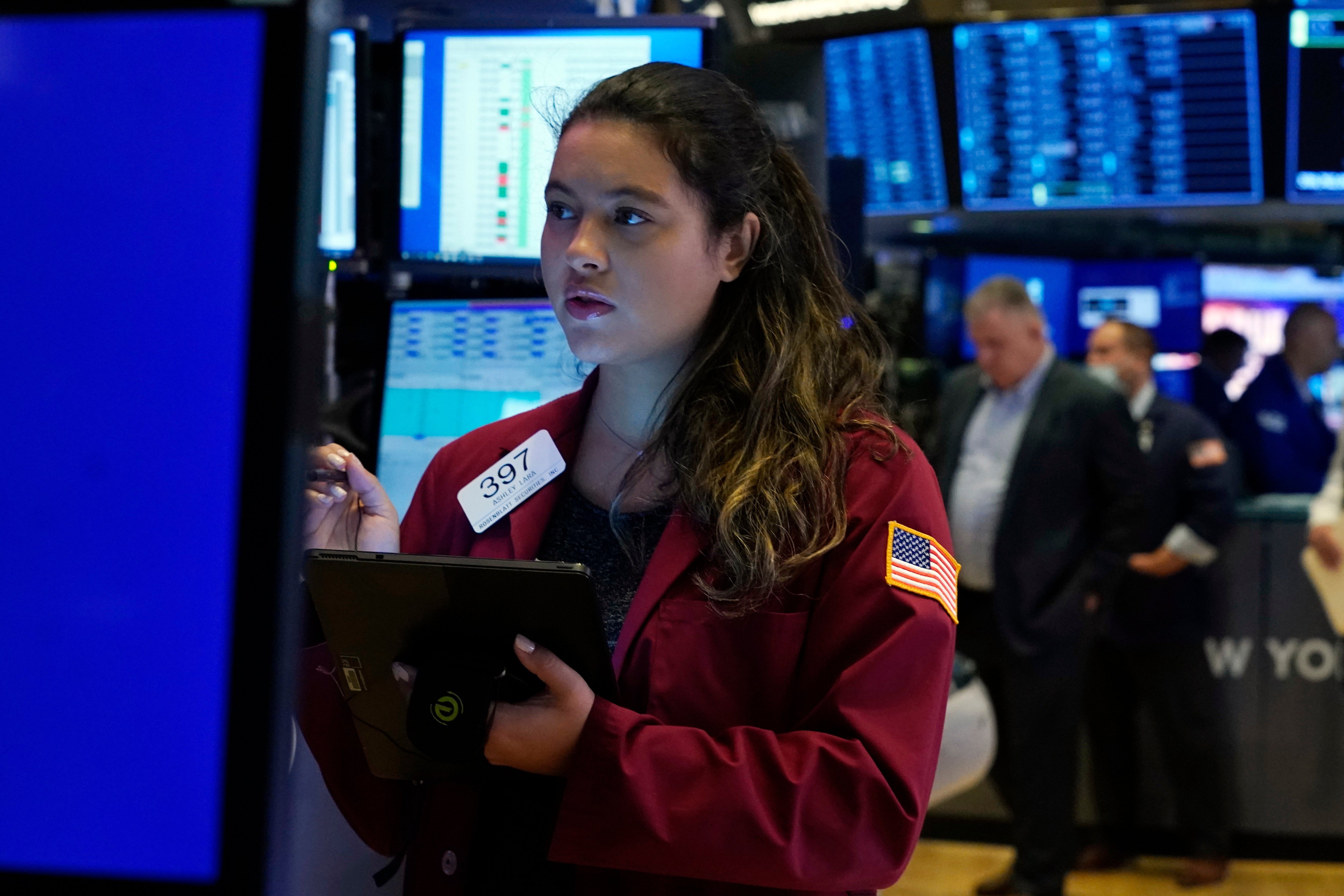 Kvinnlig handlare på börsgolvet i röd jacka som håller en surfplatta framför blå handelsskärmar.