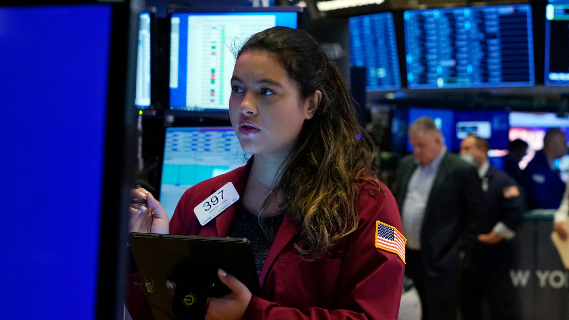 Kvinnlig handlare på börsgolvet i röd jacka som håller en surfplatta framför blå handelsskärmar.