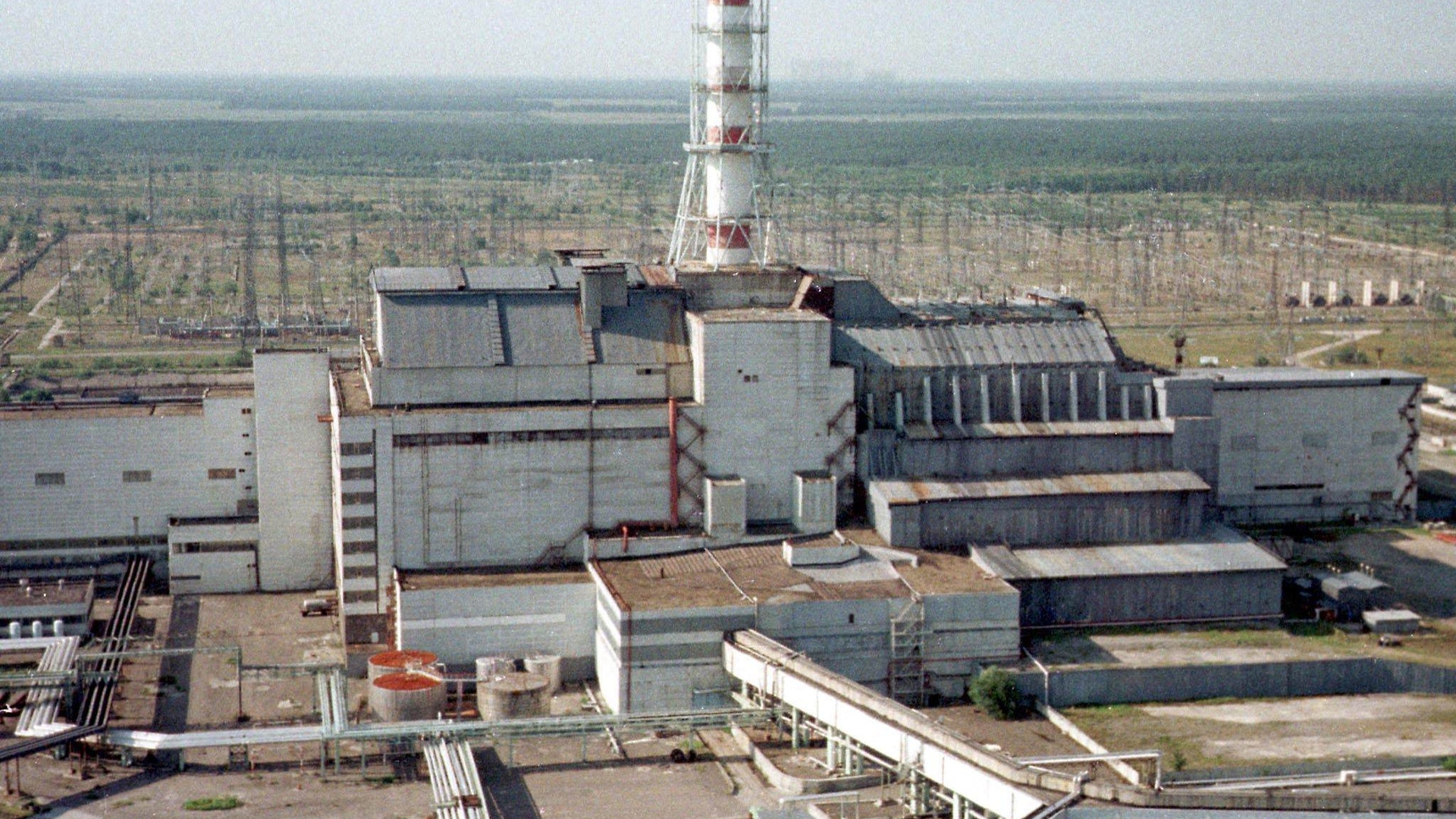 Luftfoto av Tjernobyl-kärnkraftverket med reaktorbyggnaden och kyltornet omgivet av platt, gles terräng.