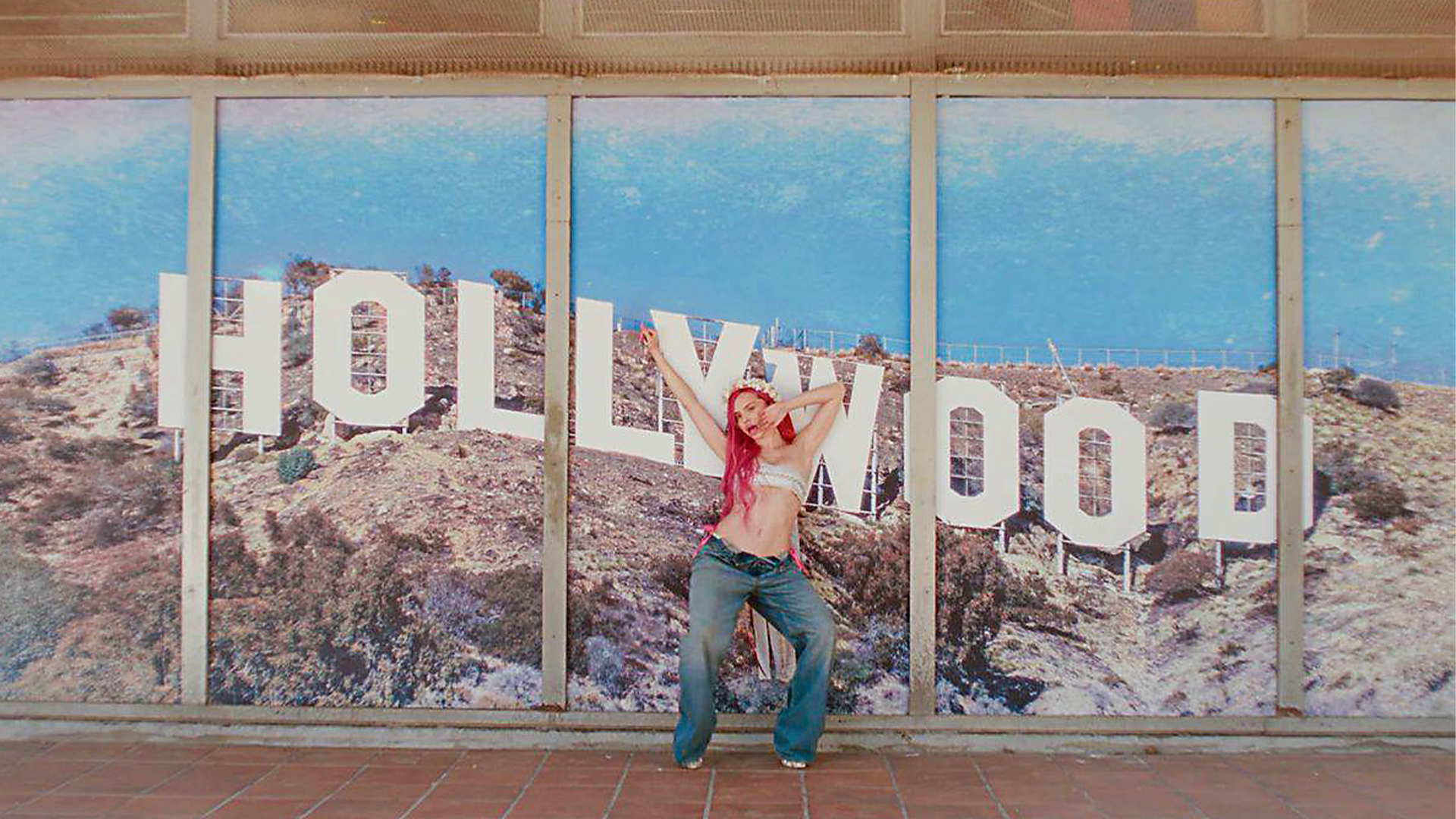 En person poserar framför ett stort fönster med Hollywood-skyltningen och Hollywood Hills synlig i bakgrunden.