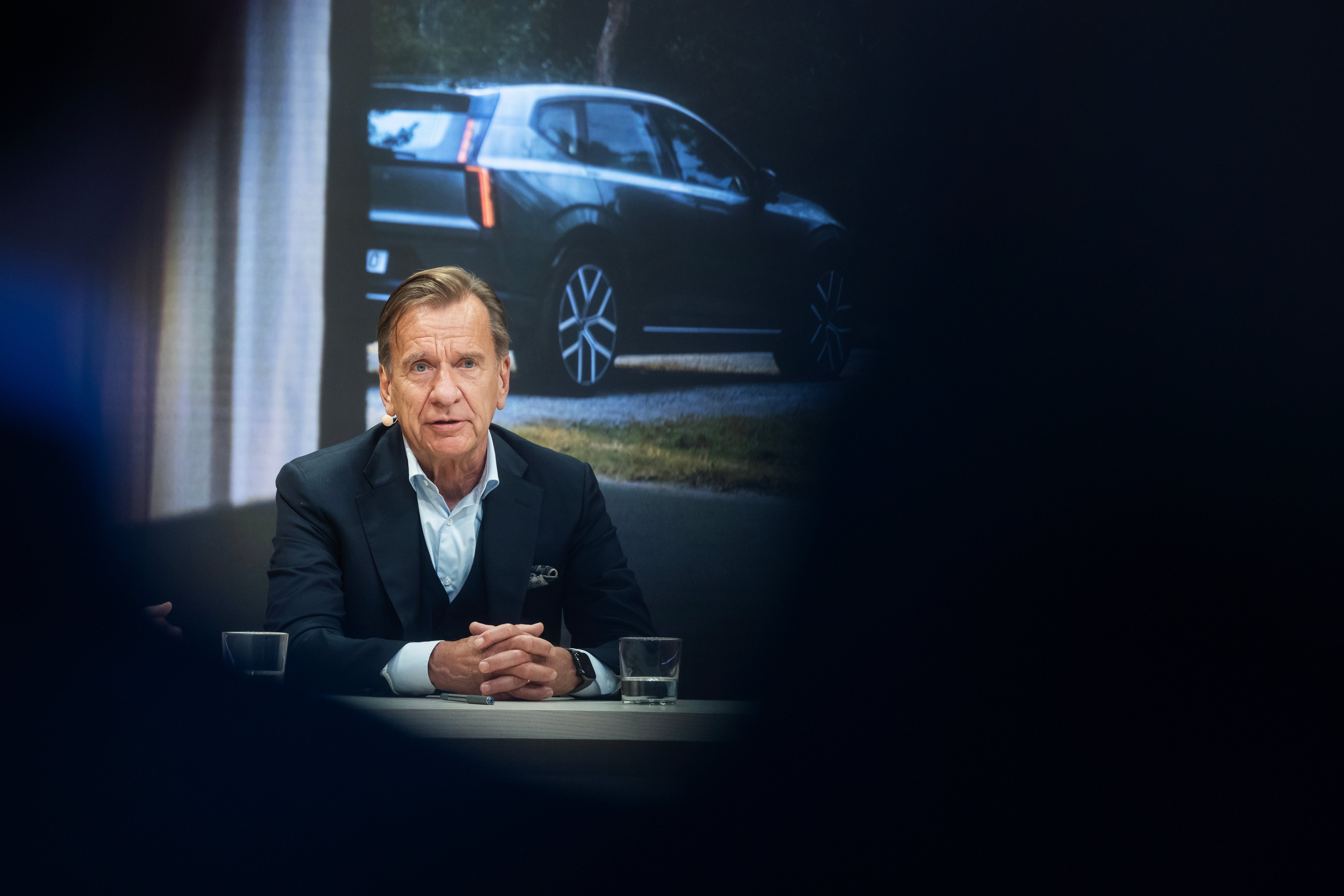 En man i kostym sitter vid ett bord under en presskonferens, med en bild av en Volvo-bil projicerad på skärmen bakom honom.