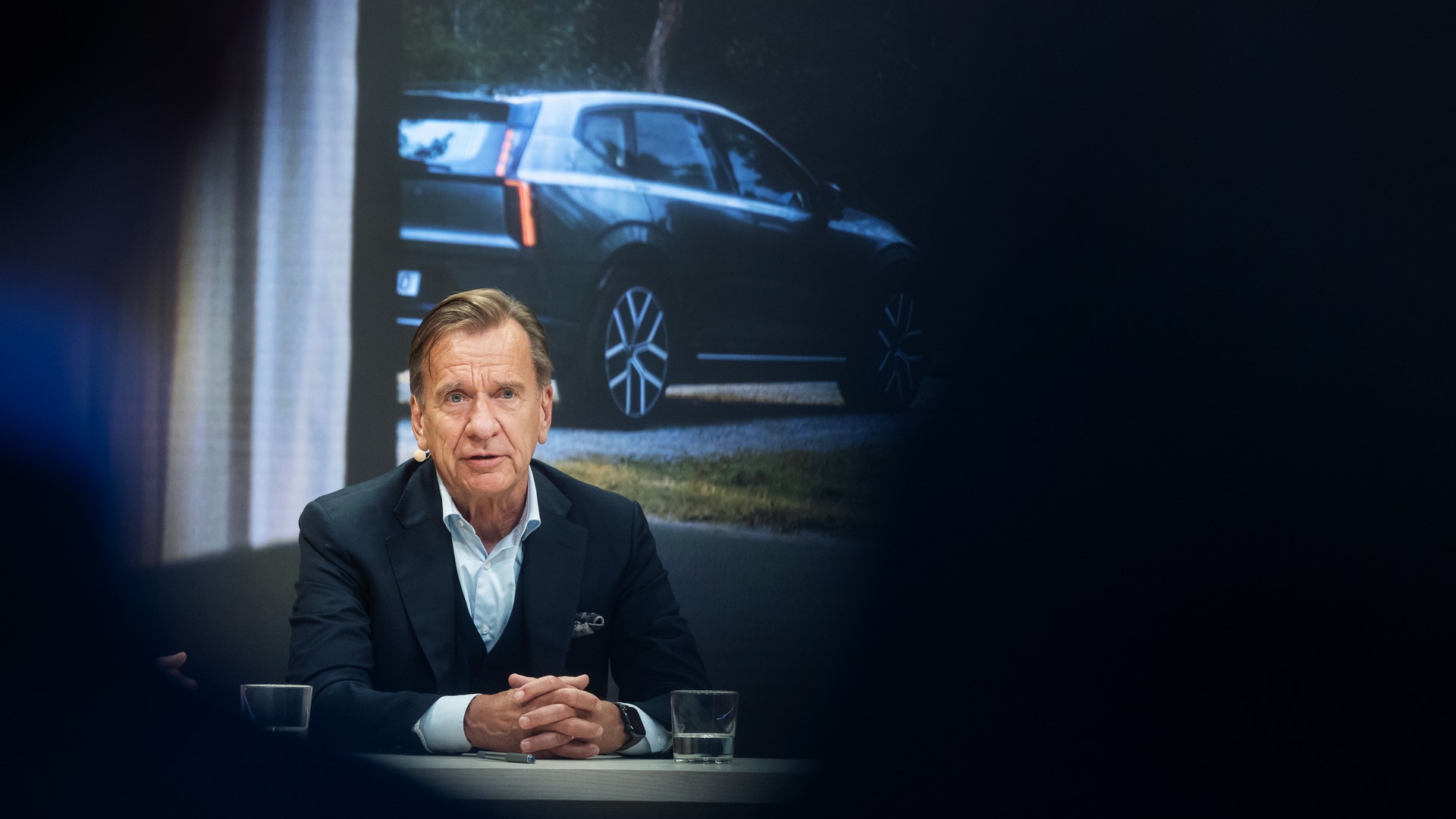 En man i kostym sitter vid ett bord under en presskonferens, med en bild av en Volvo-bil projicerad på skärmen bakom honom.