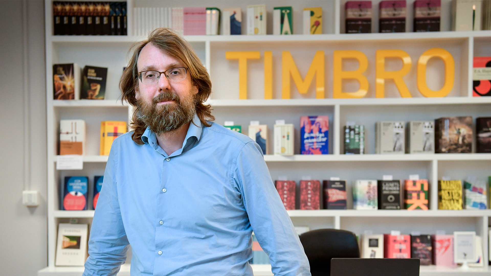 Man med långt hår och skägg i ljusblå skjorta framför en bokhylla med gula TIMBRO-bokstäver på väggen bakom sig.