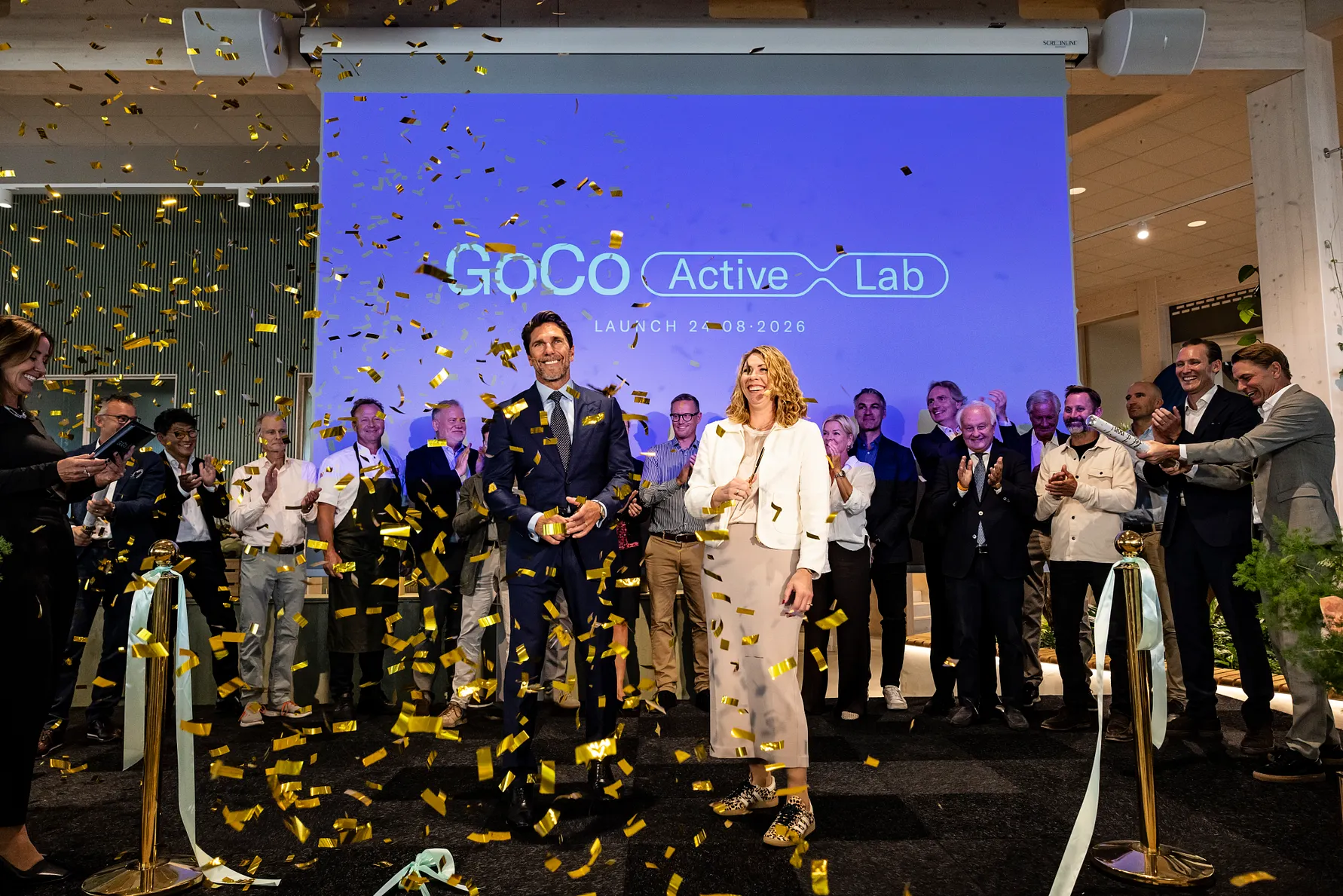 En grupp människor på en lansering av GoCo Active Lab. Guldkonfetti faller från luften medan deltagare i formell klädsel står framför en blå skärm med logotypen för GoCo Active Lab. Det finns cyklar på scenen och en festlig atmosfär.