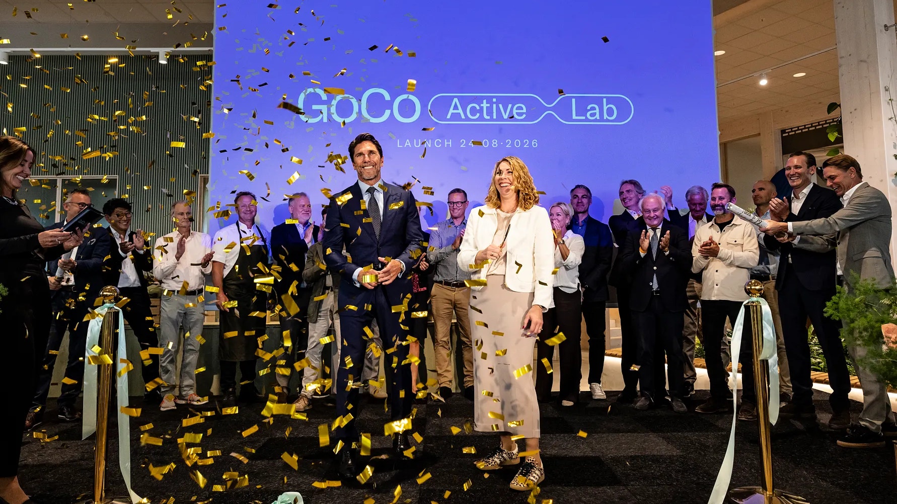 En grupp människor på en lansering av GoCo Active Lab. Guldkonfetti faller från luften medan deltagare i formell klädsel står framför en blå skärm med logotypen för GoCo Active Lab. Det finns cyklar på scenen och en festlig atmosfär.