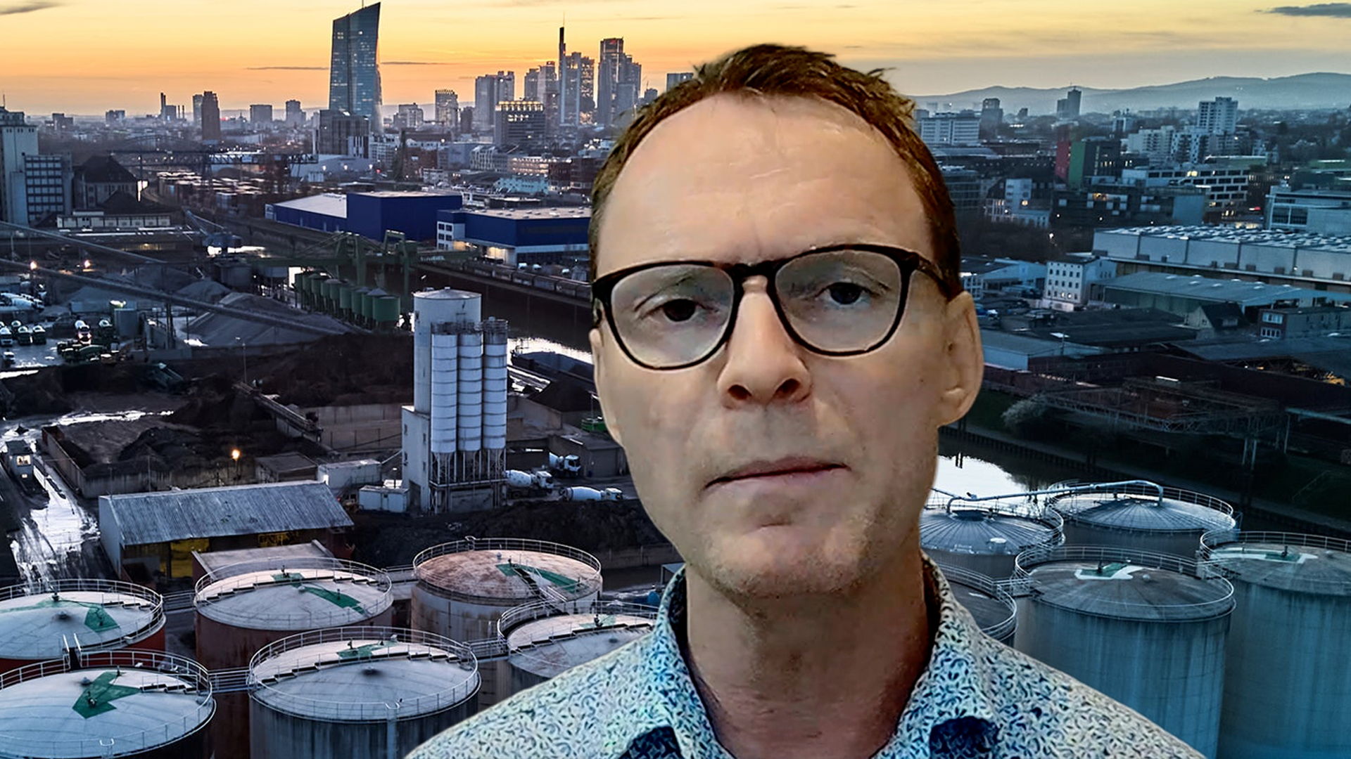 Man med glasögon framför en stadshorisont med höghus och industrianläggningar vid solnedgång.