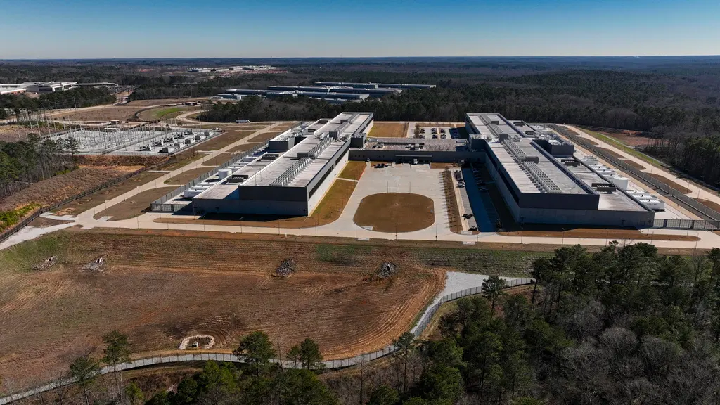 Flygfoto över en stor industrianläggning med vita byggnader omgiven av skog och öppen mark.
