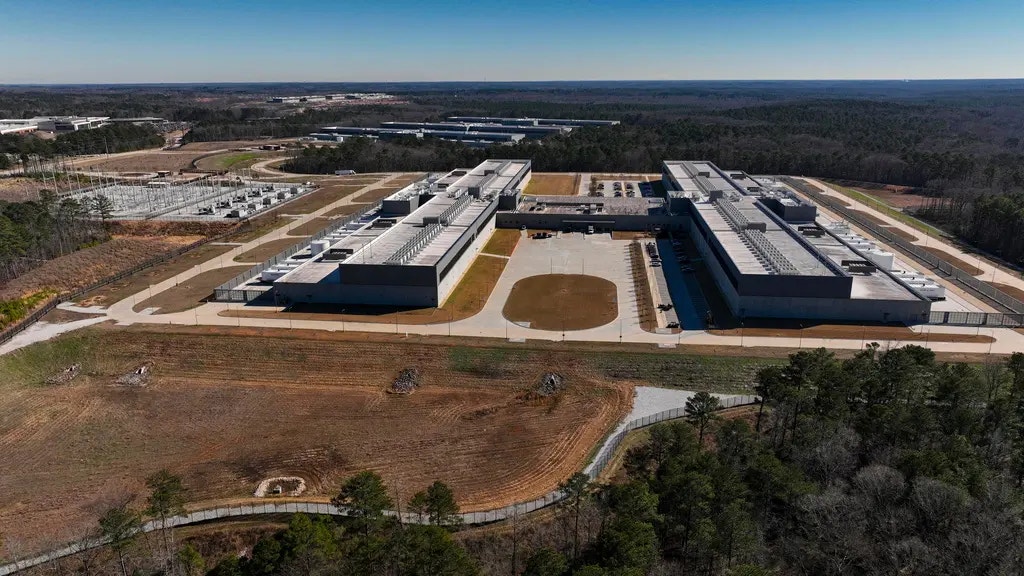 Flygfoto över en stor industrianläggning med vita byggnader omgiven av skog och öppen mark.