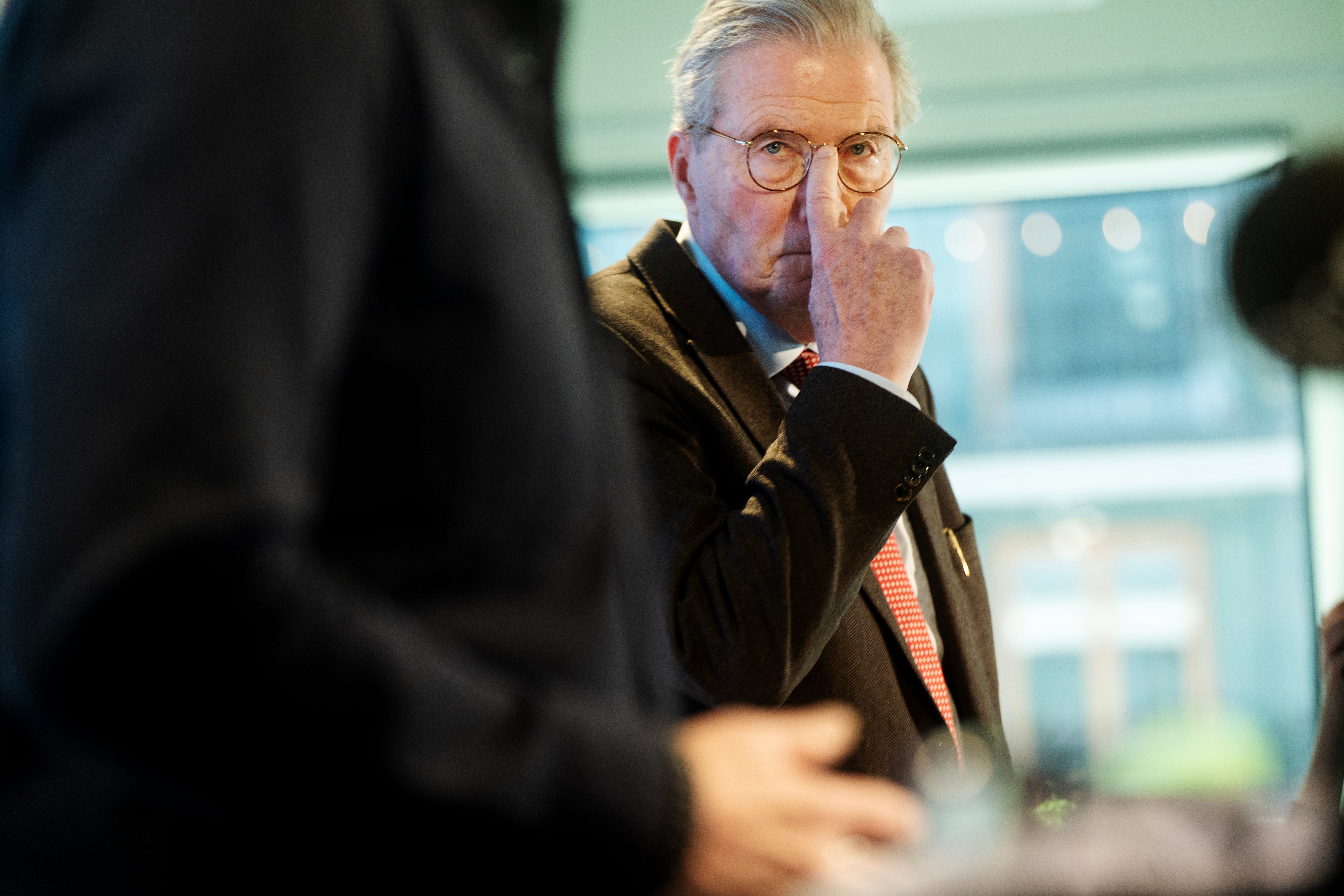 Man med glasögon i mörkblå jacka och orange slips ser tankfull ut under ett möte eller event.