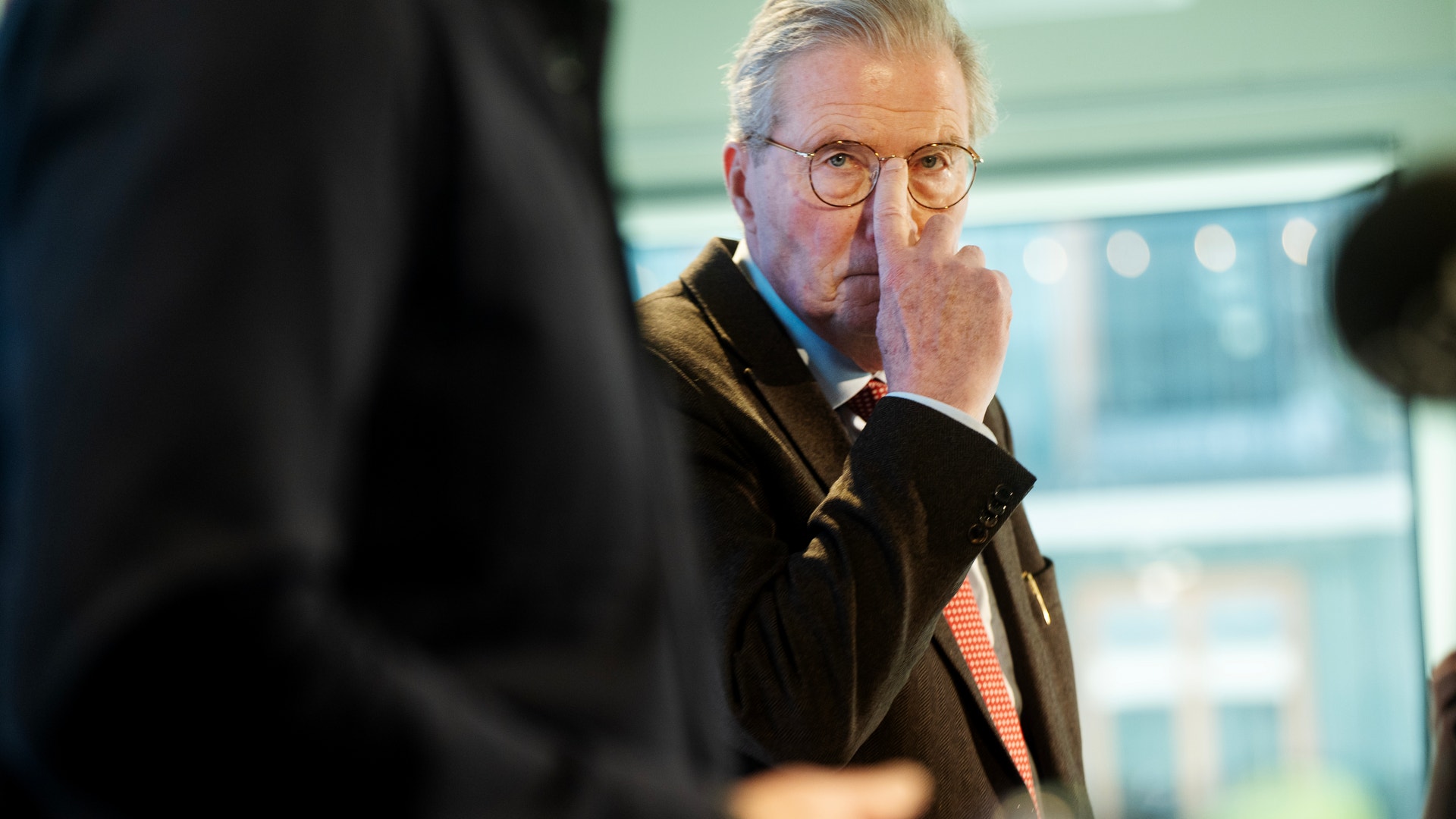 Man med glasögon i mörkblå jacka och orange slips ser tankfull ut under ett möte eller event.