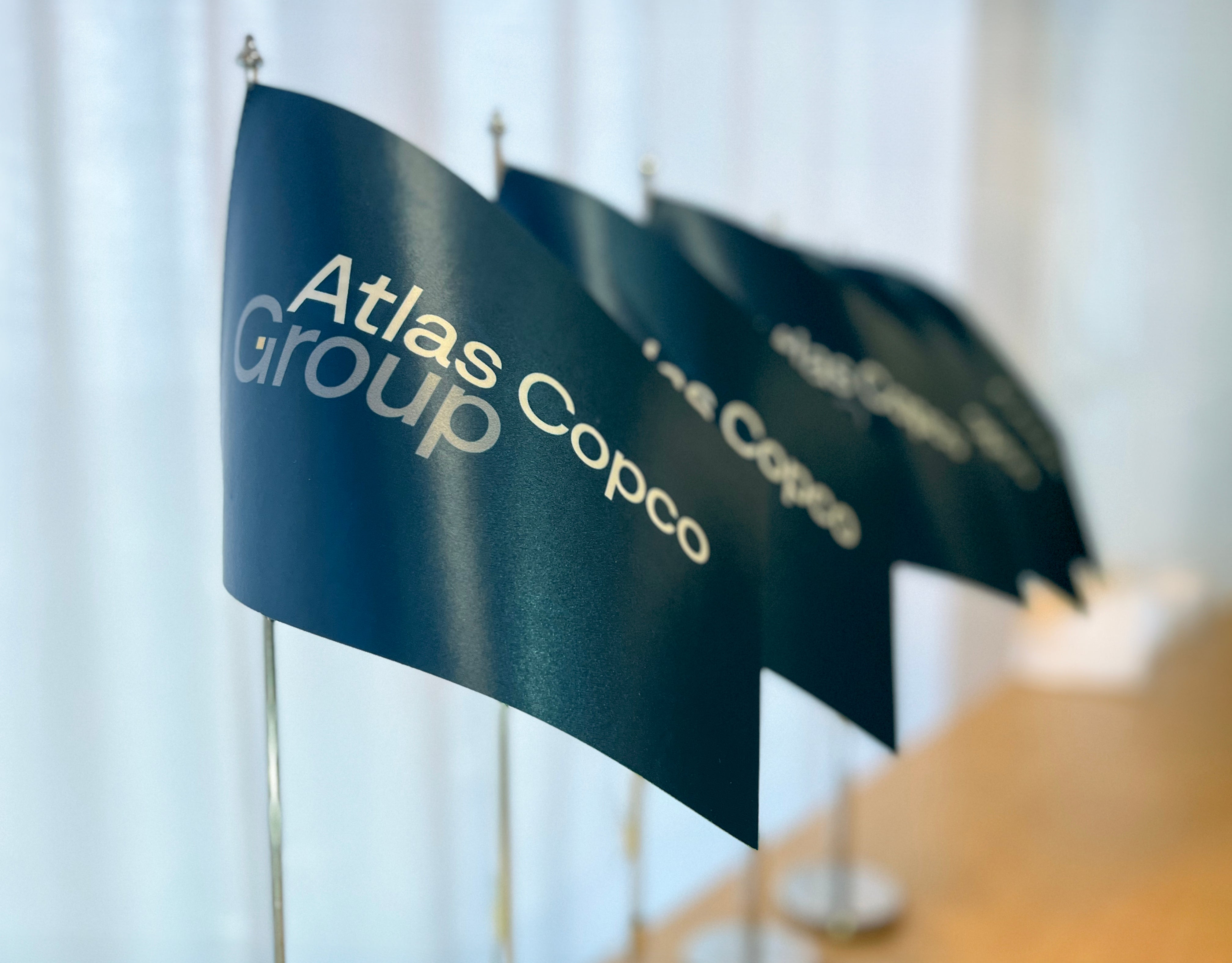 Flera blågröna flaggor med Atlas Copco Group-logotypen placerade i rad på ett skrivbord.