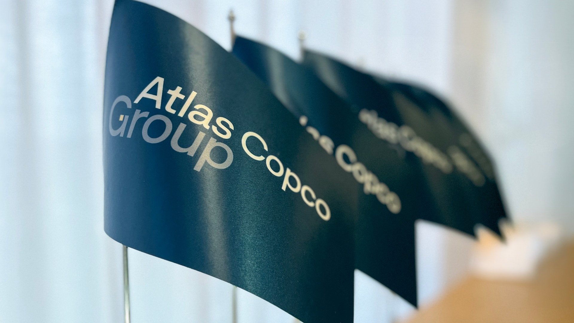 Flera blågröna flaggor med Atlas Copco Group-logotypen placerade i rad på ett skrivbord.