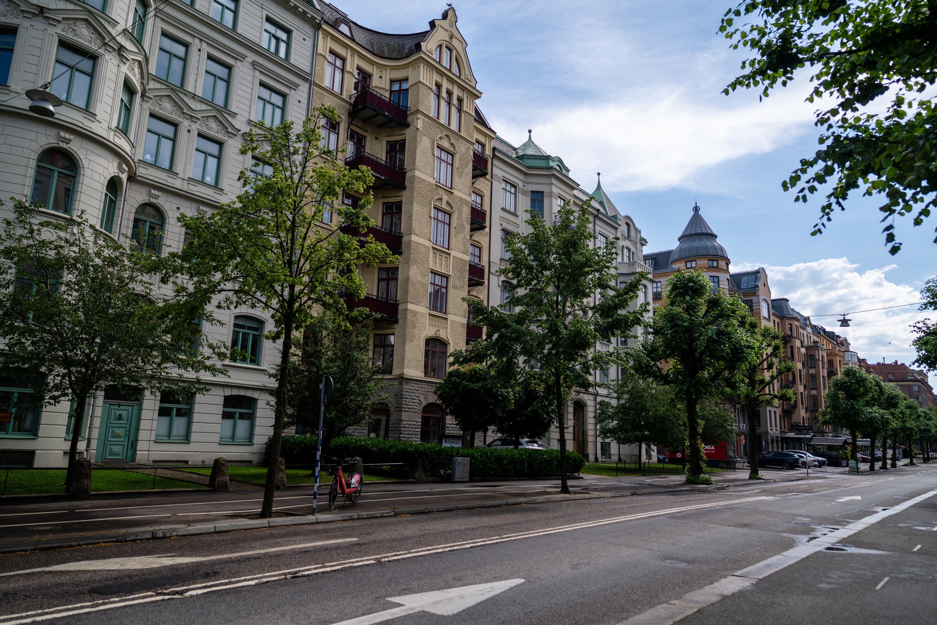 En bred gata med klassiska flervåningshus i gul och röd sten. Träd planterade längs vägen, blå himmel med moln, och några människor på gatan.