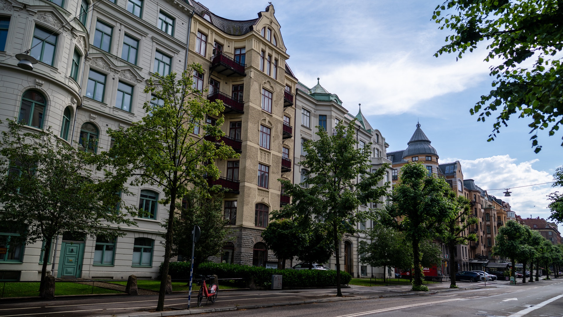 En bred gata med klassiska flervåningshus i gul och röd sten. Träd planterade längs vägen, blå himmel med moln, och några människor på gatan.