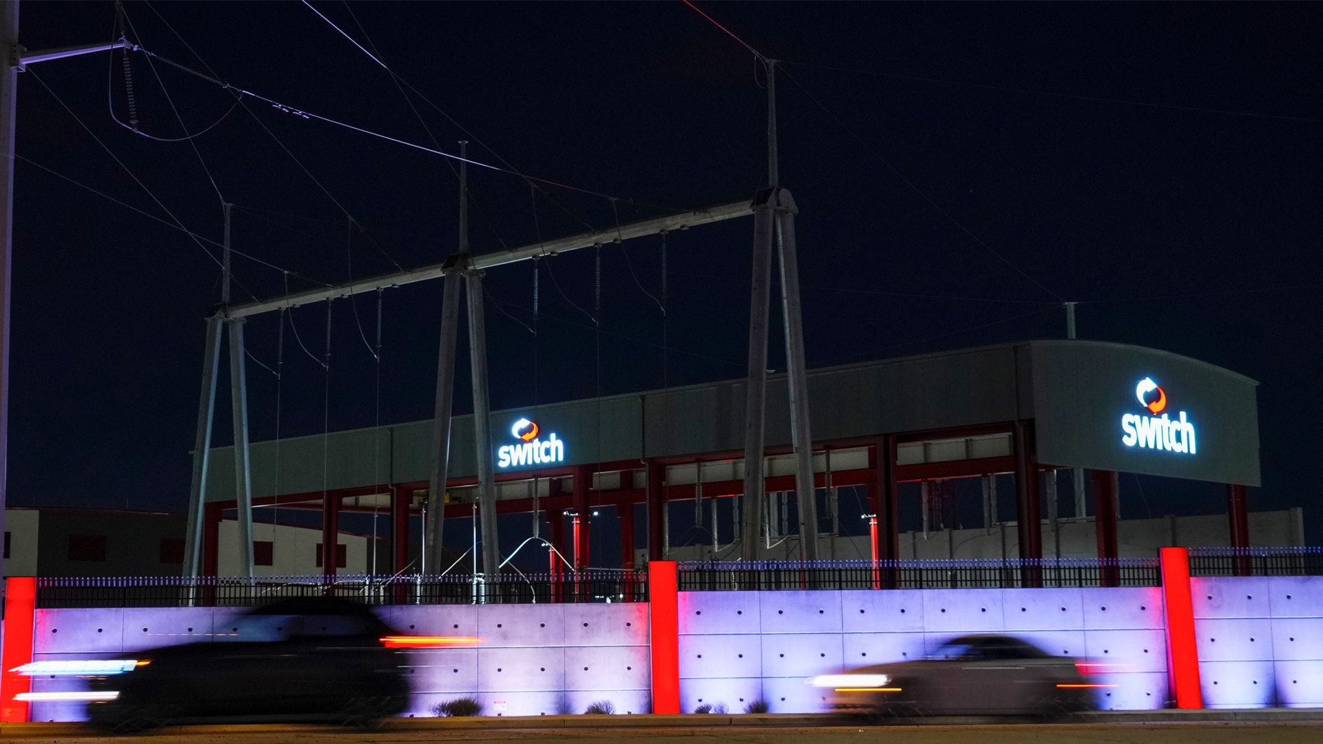 En modern byggnad fotograferad på kvällen med lila och röd neonbelysning. Byggnaden har Switch-skylt och är omgiven av belyst infrastruktur.
