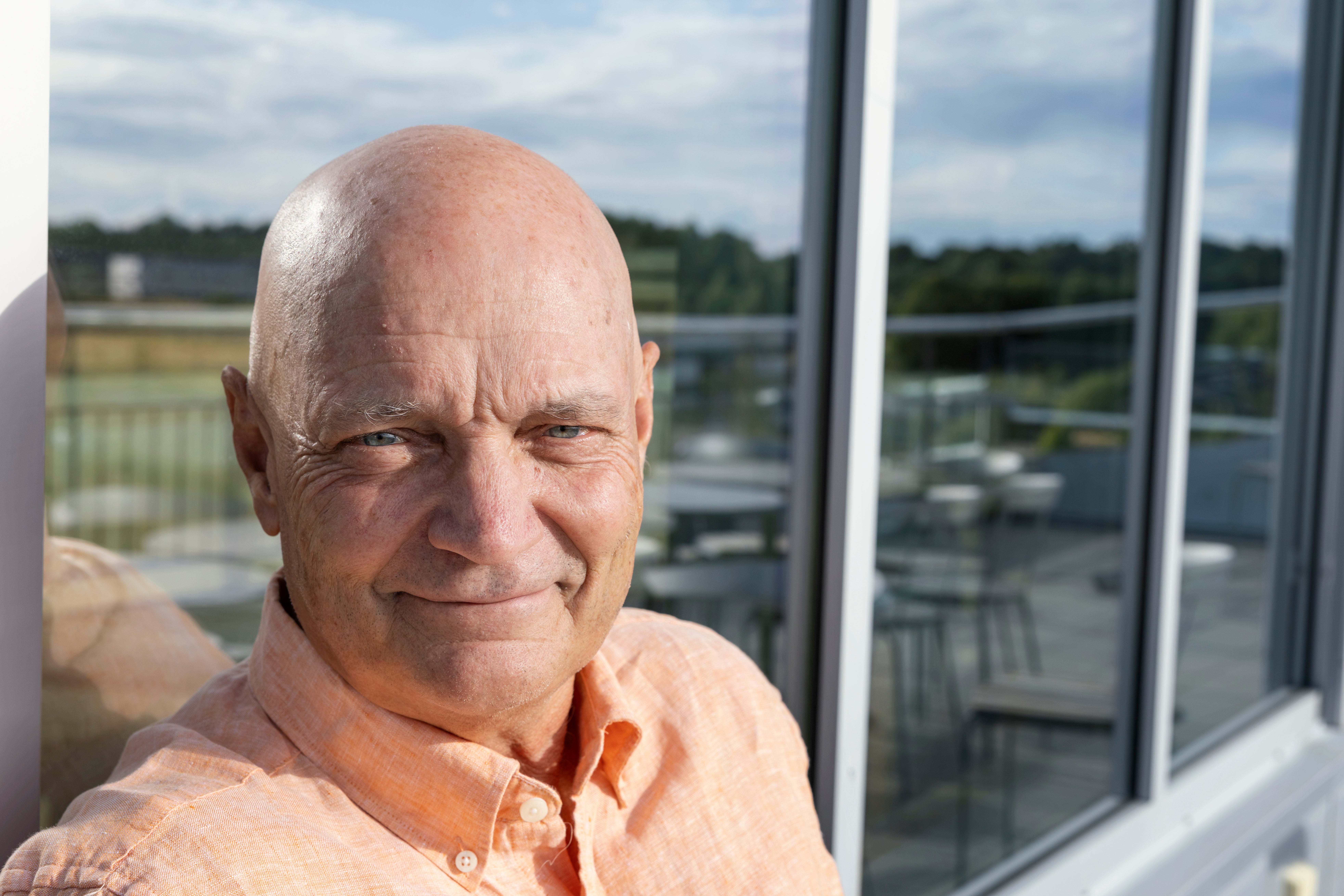 Porträtt av en man i orange skjorta som ler mot kameran. Han står framför stora fönster med utsikt över ett landskap med gröna fält.