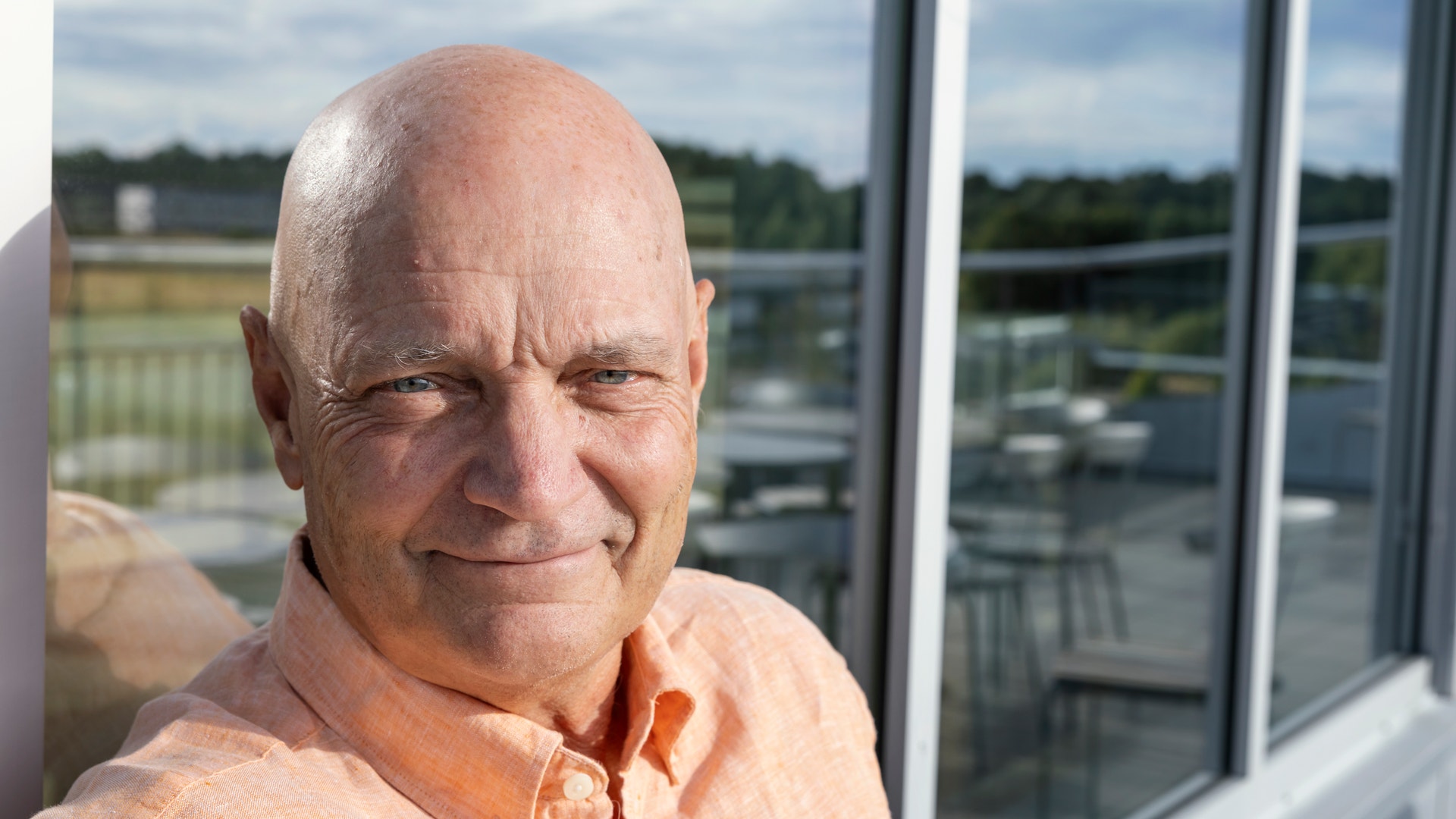 Porträtt av en man i orange skjorta som ler mot kameran. Han står framför stora fönster med utsikt över ett landskap med gröna fält.