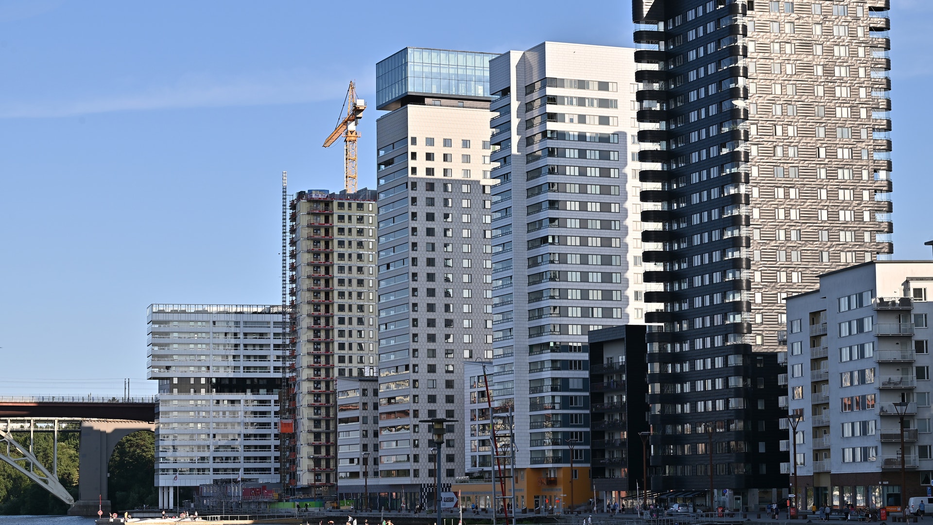 Moderna höghus längs en vattenkant under blå himmel. Flera bostadsbyggnader i olika höjder och färger, från vita till mörka fasader, med en byggkran synlig mellan husen.