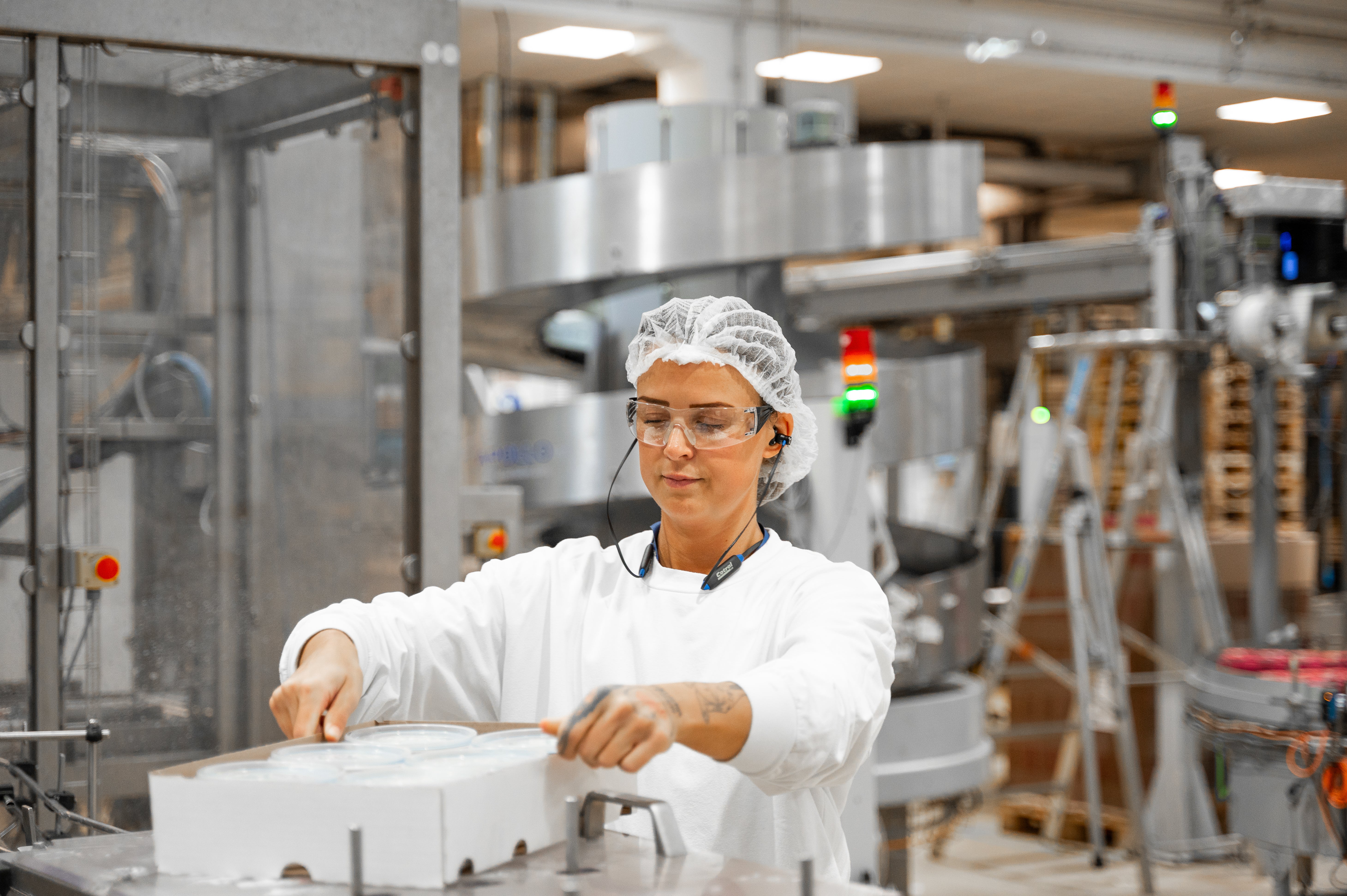 Medarbetare i vita arbetskläder och hårnät arbetar vid en maskin i en mejeriproduktion.