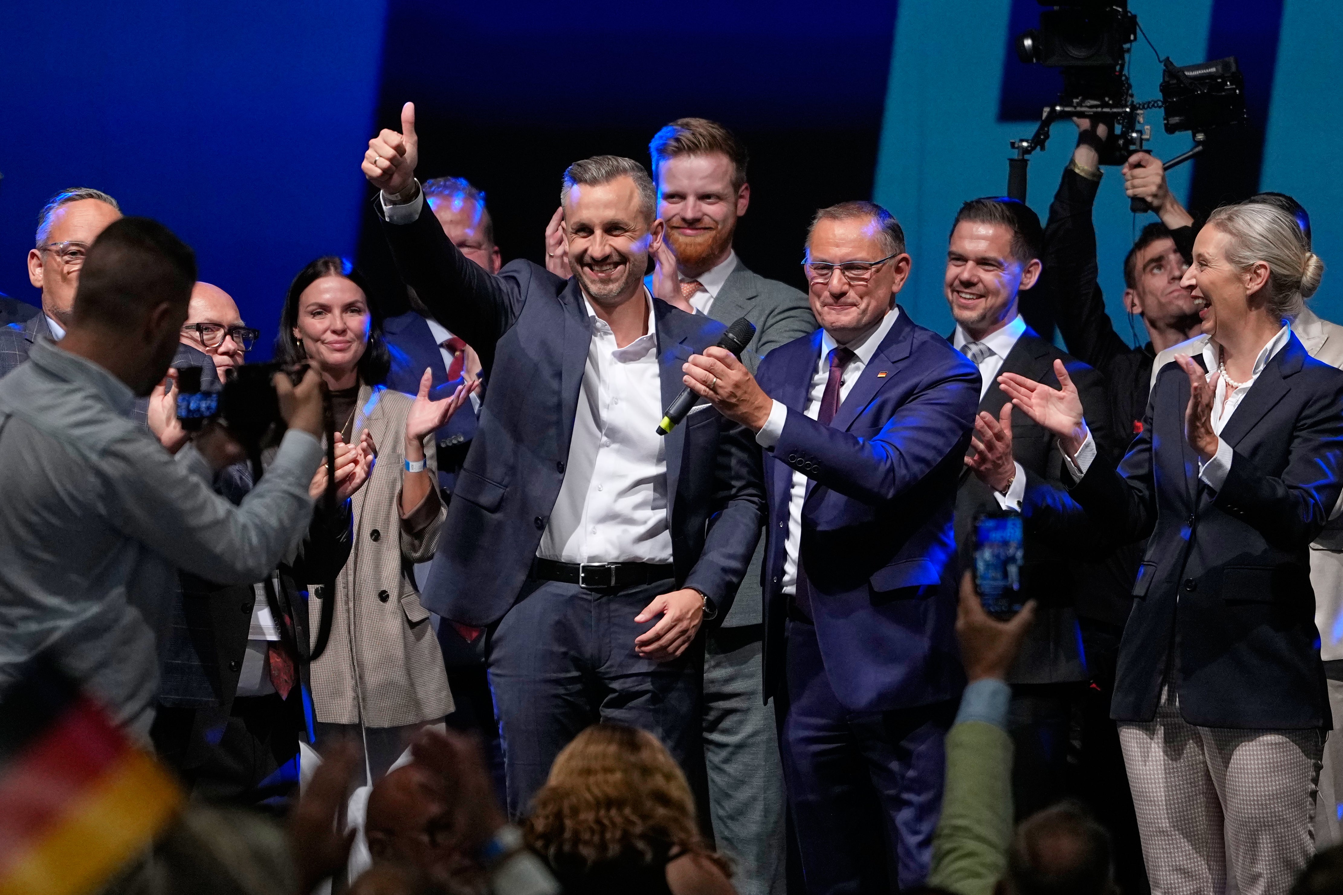 En grupp människor i formell klädsel står tillsammans på en scen med blå belysning. En man i mitten gör en triumfgest med tummen upp medan gruppen applåderar.