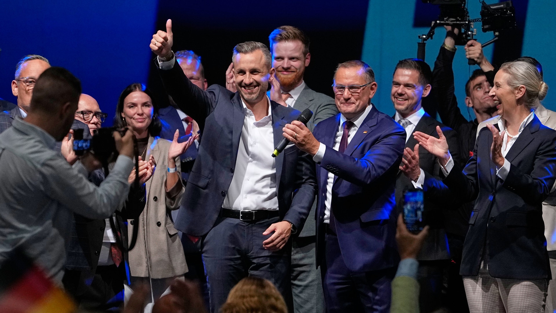 En grupp människor i formell klädsel står tillsammans på en scen med blå belysning. En man i mitten gör en triumfgest med tummen upp medan gruppen applåderar.
