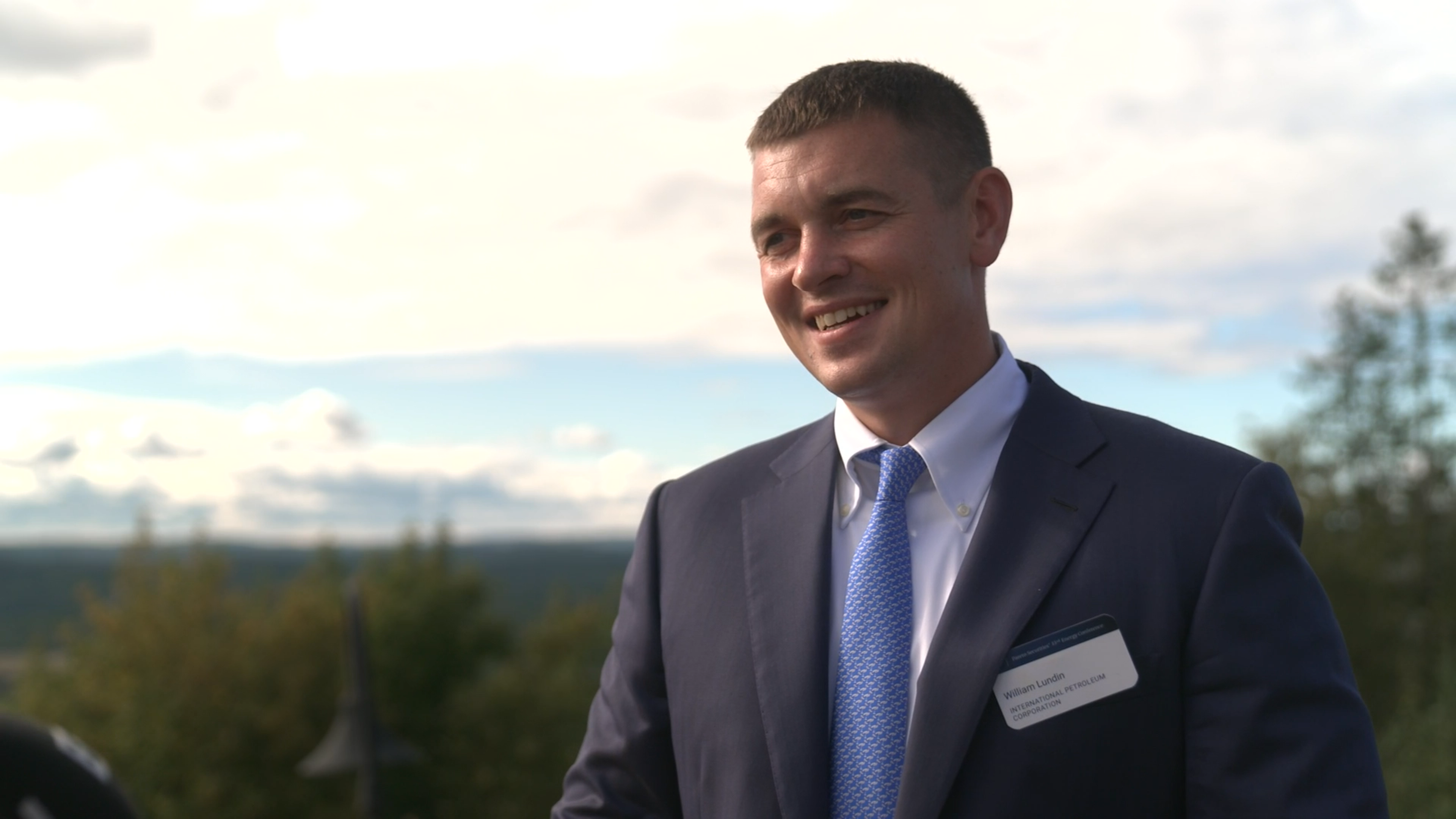 William Lundin i kostym och slips står utomhus med skog och himmel i bakgrunden.