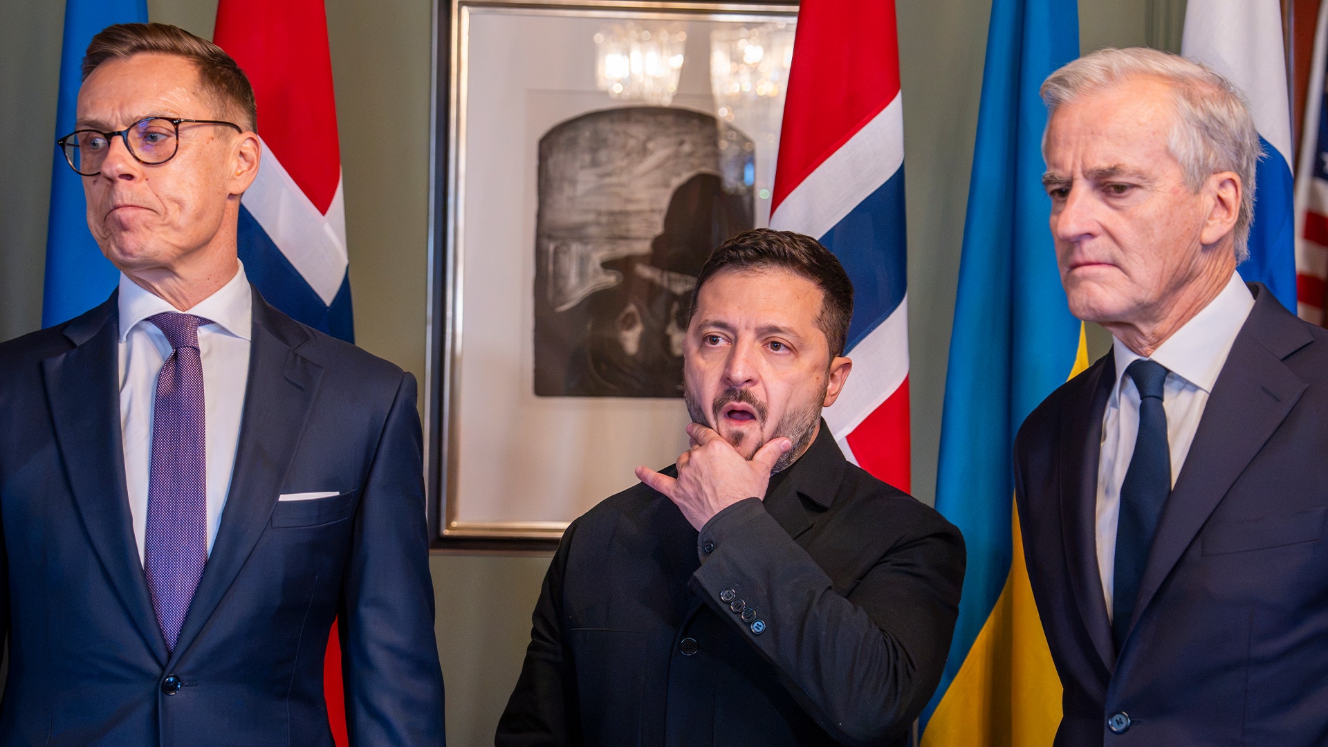 Tre män i mörka kostymer står framför flaggor. Mannen i mitten för handen mot hakan och tittar åt sidan.