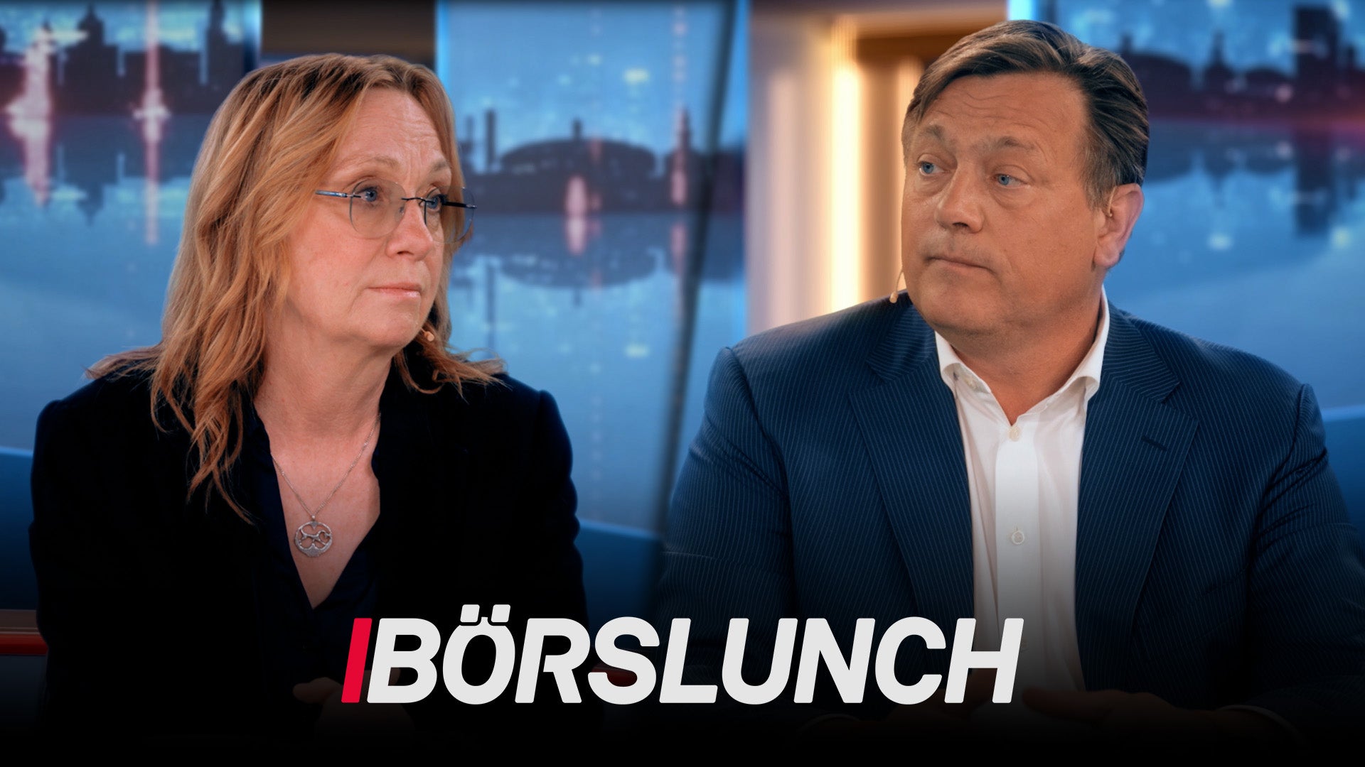 Två personer sitter i en TV-studio för programmet Börslunch. En kvinna och en man är fotograferade framför stora fönster med stadsvy i bakgrunden.