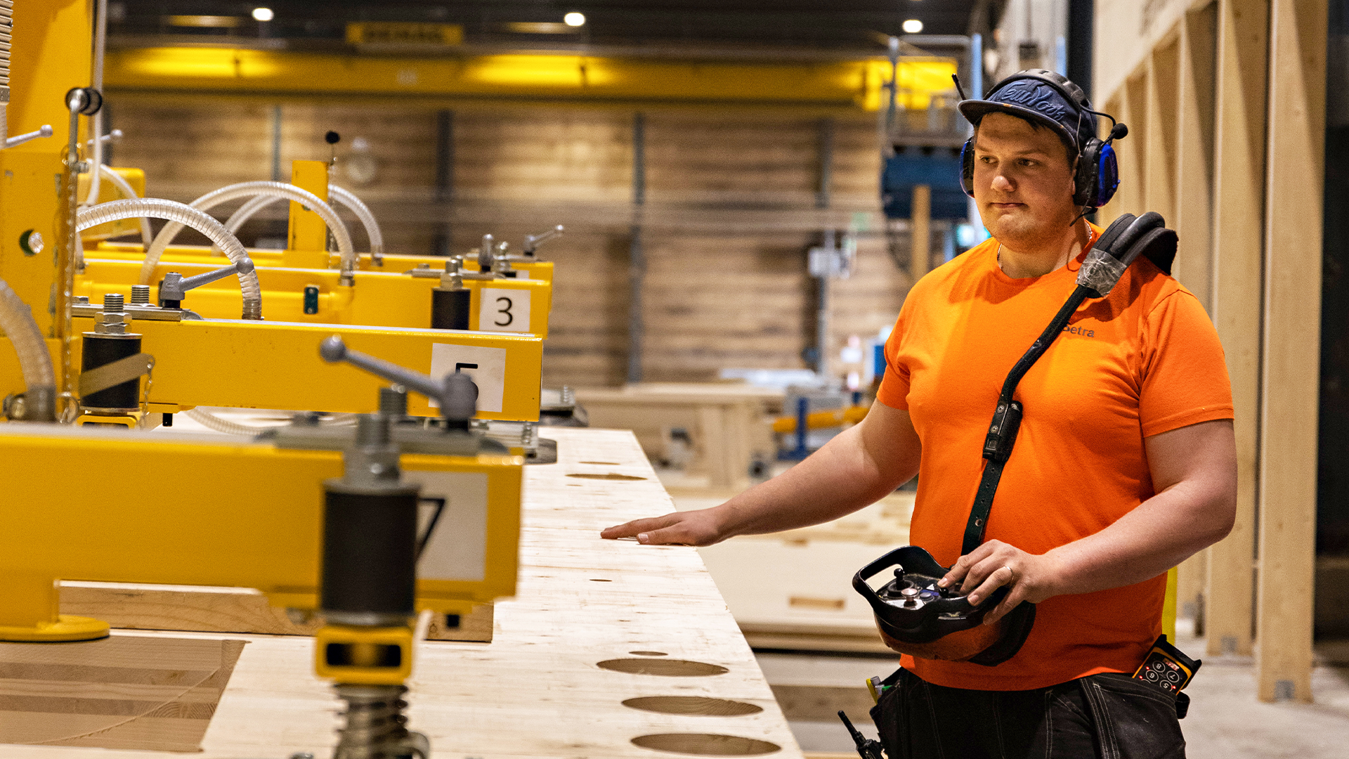 En man i orange t-shirt, hörselskydd och keps står i en träfabrik och styr en gul maskin med en handhållen fjärrkontroll.