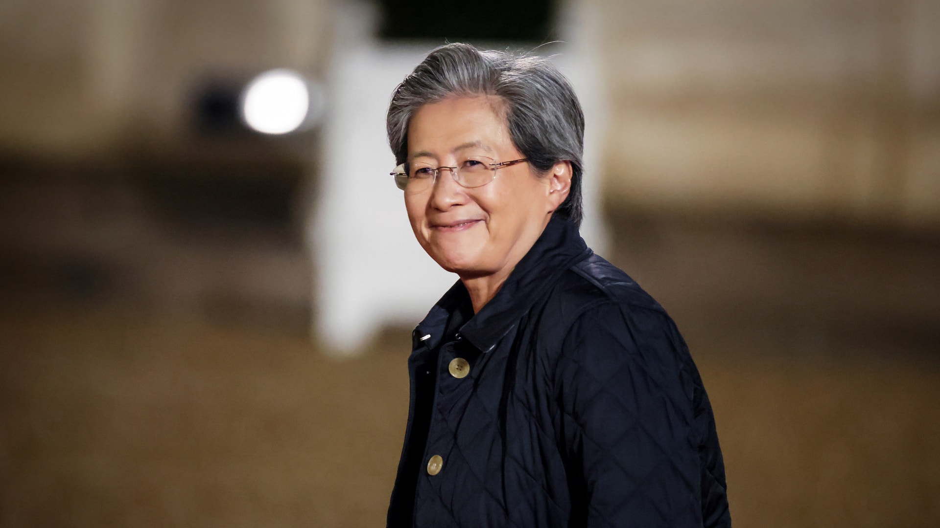 Porträtt av en person med glasögon och mörkblå jacka mot en suddig bakgrund inomhus.