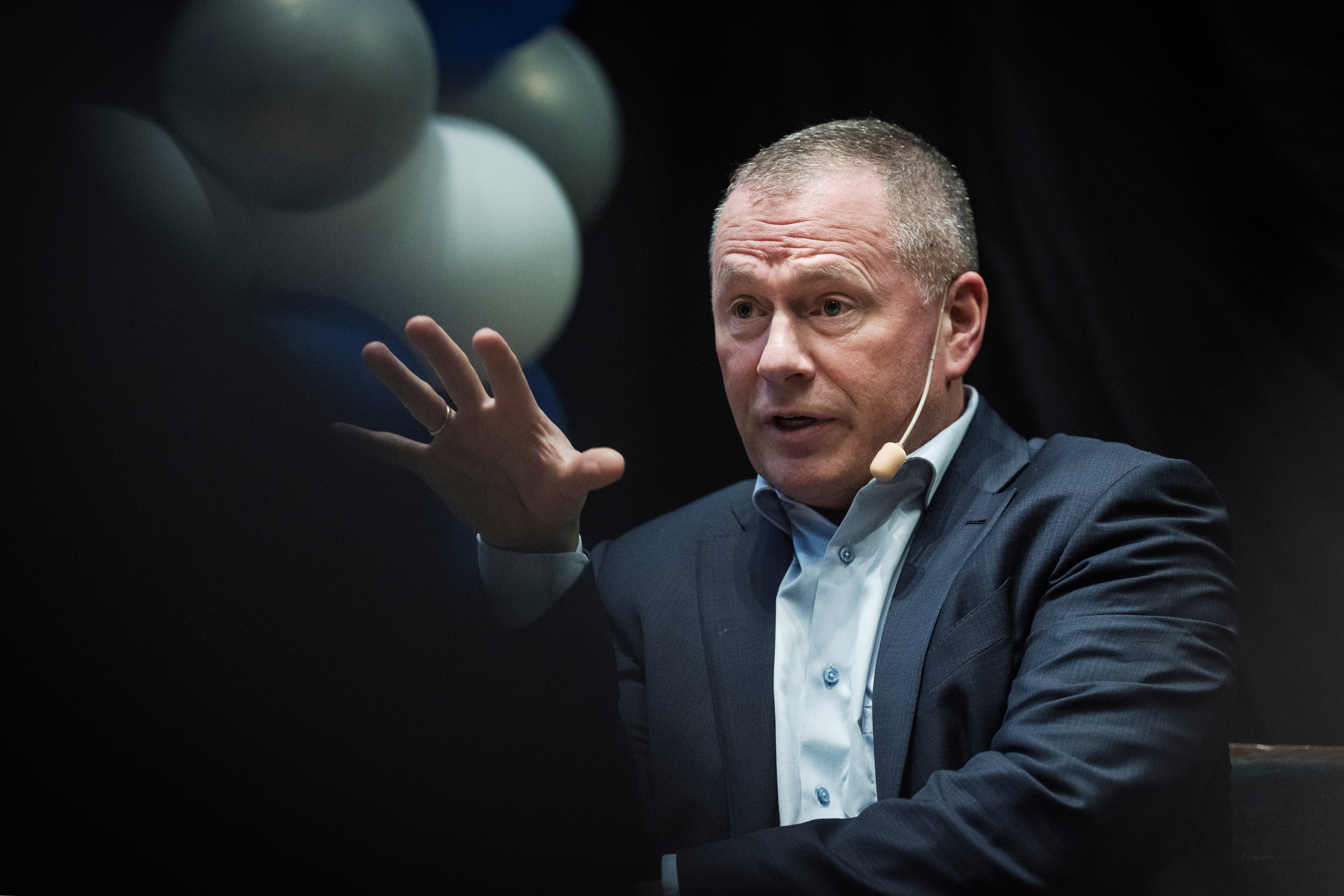 En man i mörk kostym och ljus skjorta gestikulerar medan han talar. Han har kort gråsprängt hår och presenterar framför en mörk bakgrund med ljusa sfärer.
