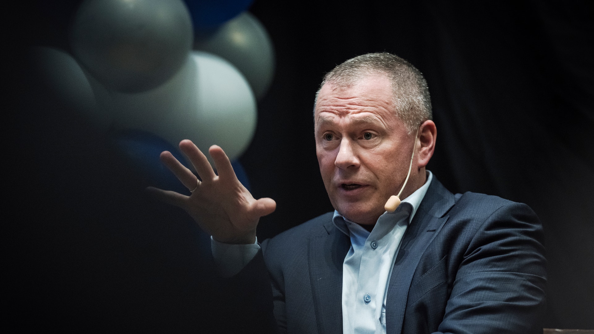 En man i mörk kostym och ljus skjorta gestikulerar medan han talar. Han har kort gråsprängt hår och presenterar framför en mörk bakgrund med ljusa sfärer.