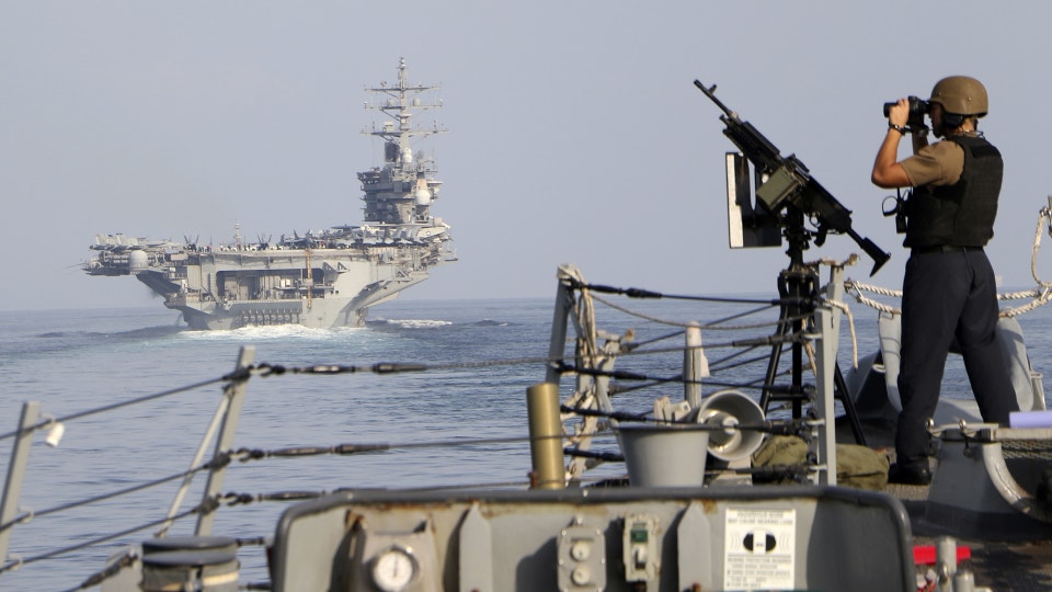 Militär soldat på ett krigsfartyg opererar ett vapen med ett hangarfartyg synligt i bakgrunden på havet.