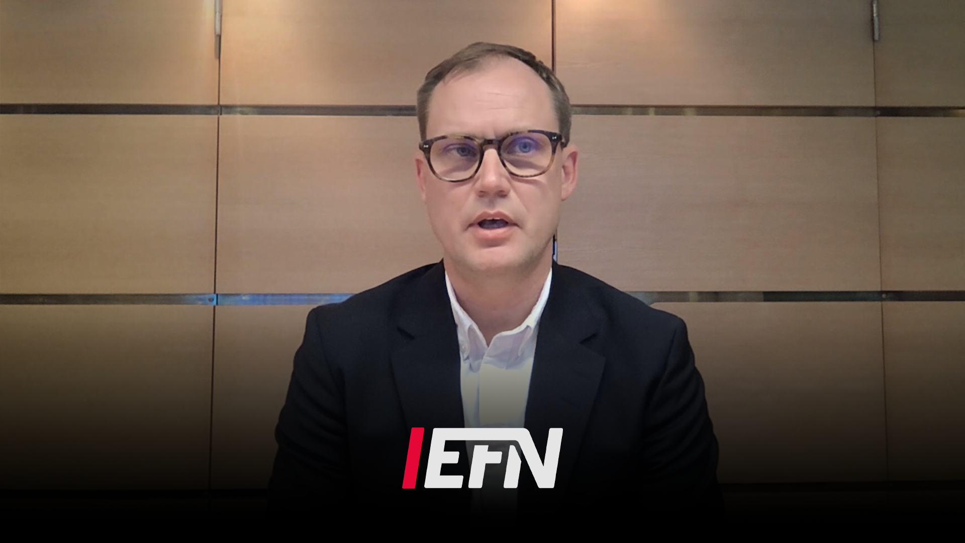Man i glasögon och mörk blazer talar framför en vägg med EFN-logotyp i en studiomiljö.