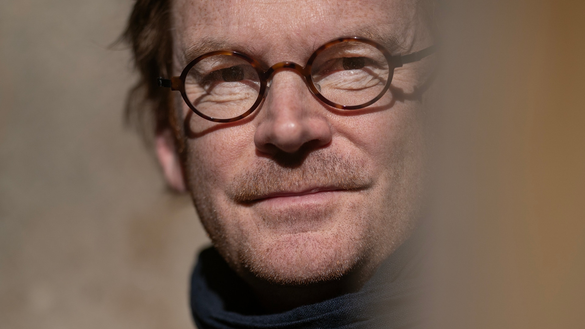 Porträtt av en man med glasögon och svart polotröja mot en neutral bakgrund.