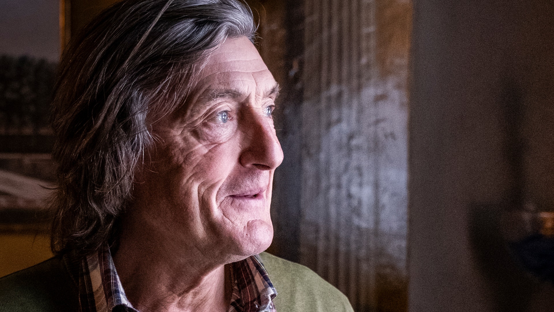 Porträtt av en äldre man i profil med långt gråvitt hår, grön tröja och ruterad skjorta.