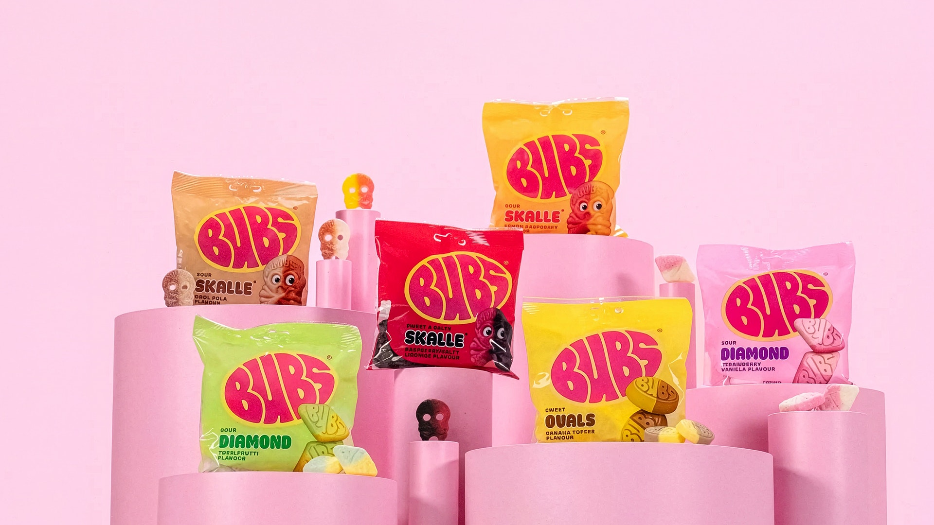 Olika paket av BIBS godis i olika färger och smaker arrangerade på rosa podier mot rosa bakgrund.