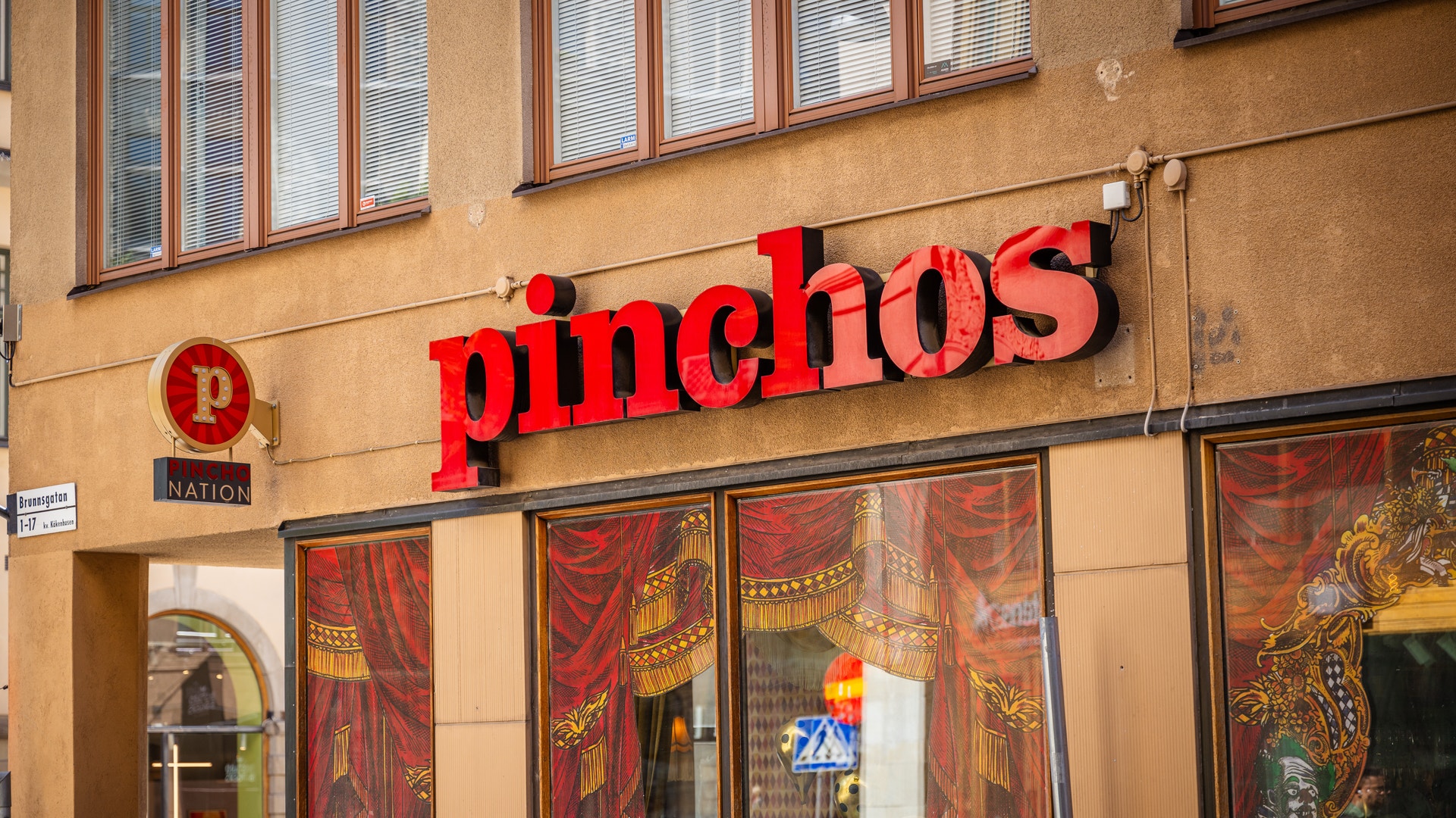 Fasaden på restaurangen Pinchos med rött skylttext på beige vägg och dekorerade röda dörrar.