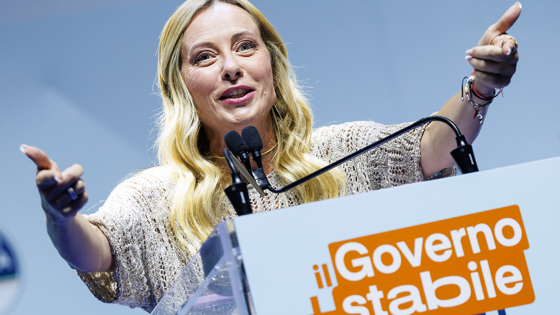 Kvinna med blont hår talar vid en mikrofon framför ett podium med texten 'Governo stabile'.