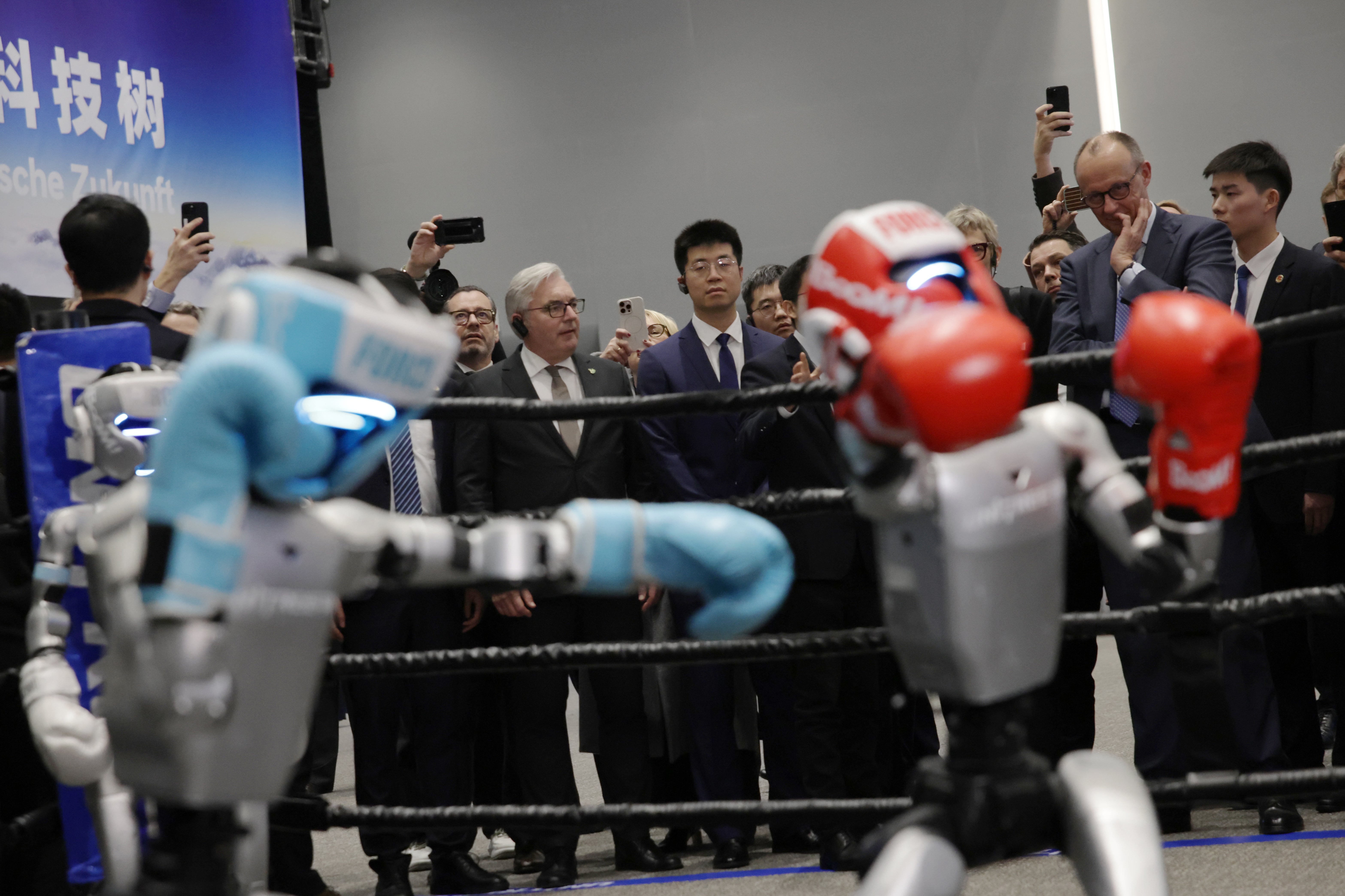 Två robotar boxar mot varandra i en ring medan en grupp människor i affärskläder står omkring och observerar med mobiltelefoner.