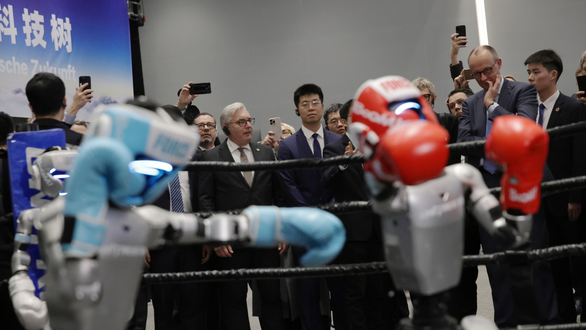 Två robotar boxar mot varandra i en ring medan en grupp människor i affärskläder står omkring och observerar med mobiltelefoner.